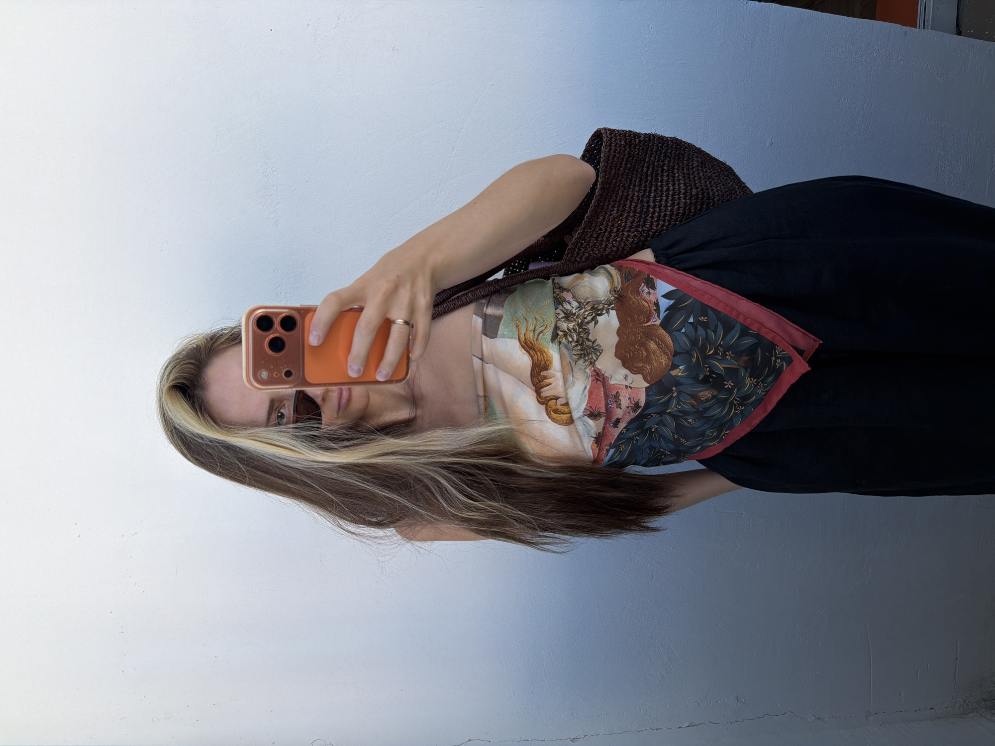 A woman is taking a selfie against a white wall. She is wearing a silk scarf as a top, black pants, sunglasses, and a gold ring. She is holding an orange smartphone in her right hand and a brown woven bag in her left. The background is a plain white wall, with a gradient of blue and white from left to right. The scene appears to be outdoors in a city, possibly in Khimki, Russia, given the architectural style of the wall and the general lighting conditions.