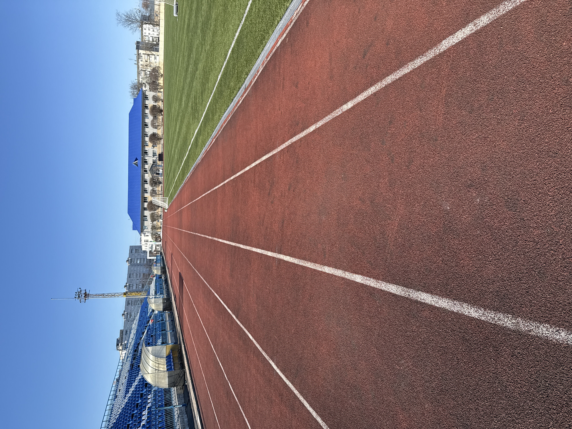 The image presents a portion of the stadium in Chaikovsky, Russia. A red running track dominates the foreground, delineated by white lane markings. To the left of the track, blue seating is visible. A lighting tower stands near the seats. Adjacent to the track, there is a green field. In the background, a blue-roofed building and other structures are seen under a clear blue sky. Bare trees are interspersed between the buildings.
