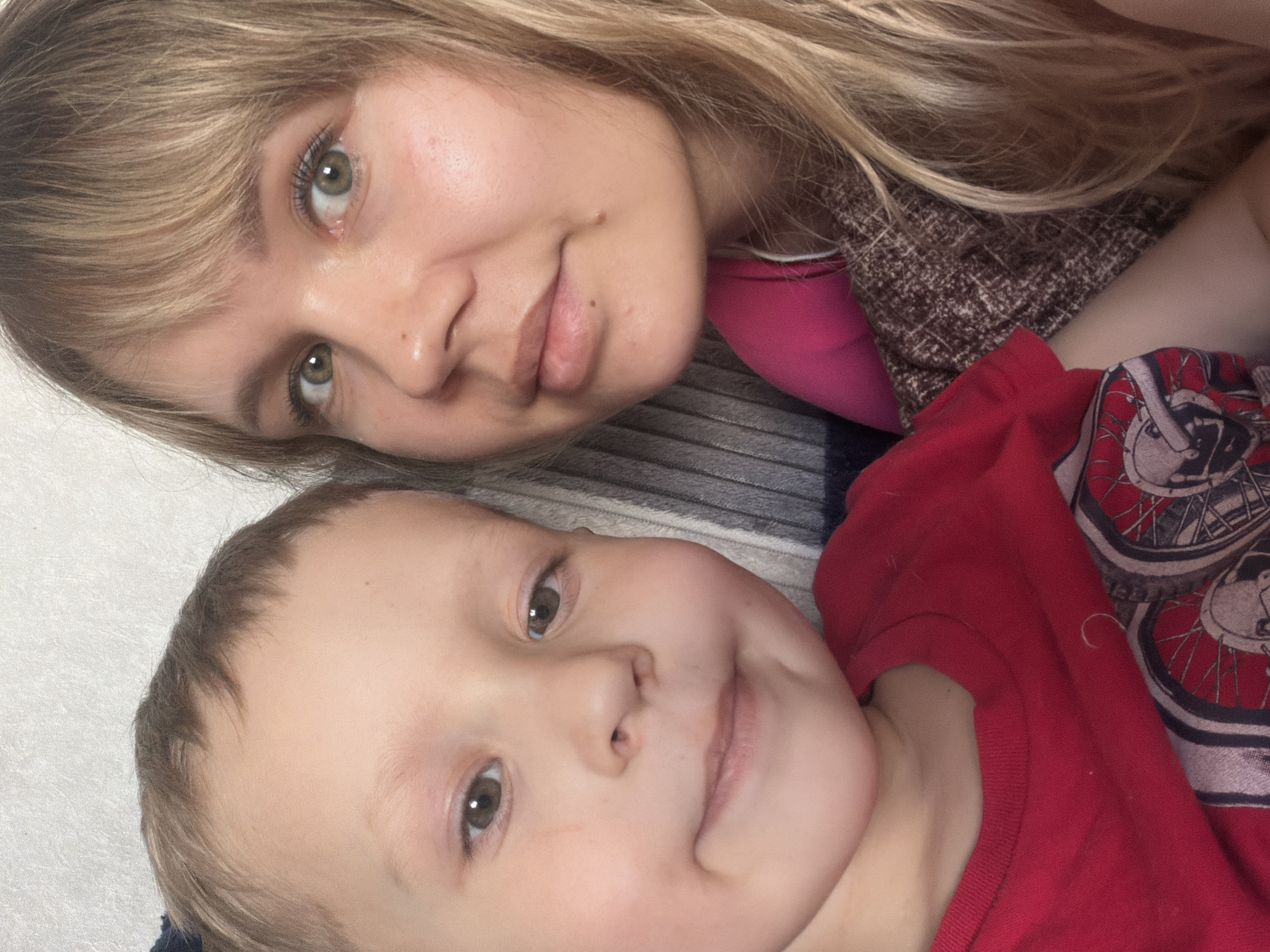 A selfie taken indoors shows two people. The person at the top of the frame is a fair-skinned female with blonde hair and green eyes. She is wearing a pink top under a brown and black patterned sweater. Below her is a fair-skinned child with short brown hair and brown eyes, wearing a red shirt with a bicycle wheel pattern. The background appears to be a gray and white striped blanket or bedsheet. The location is unknown.