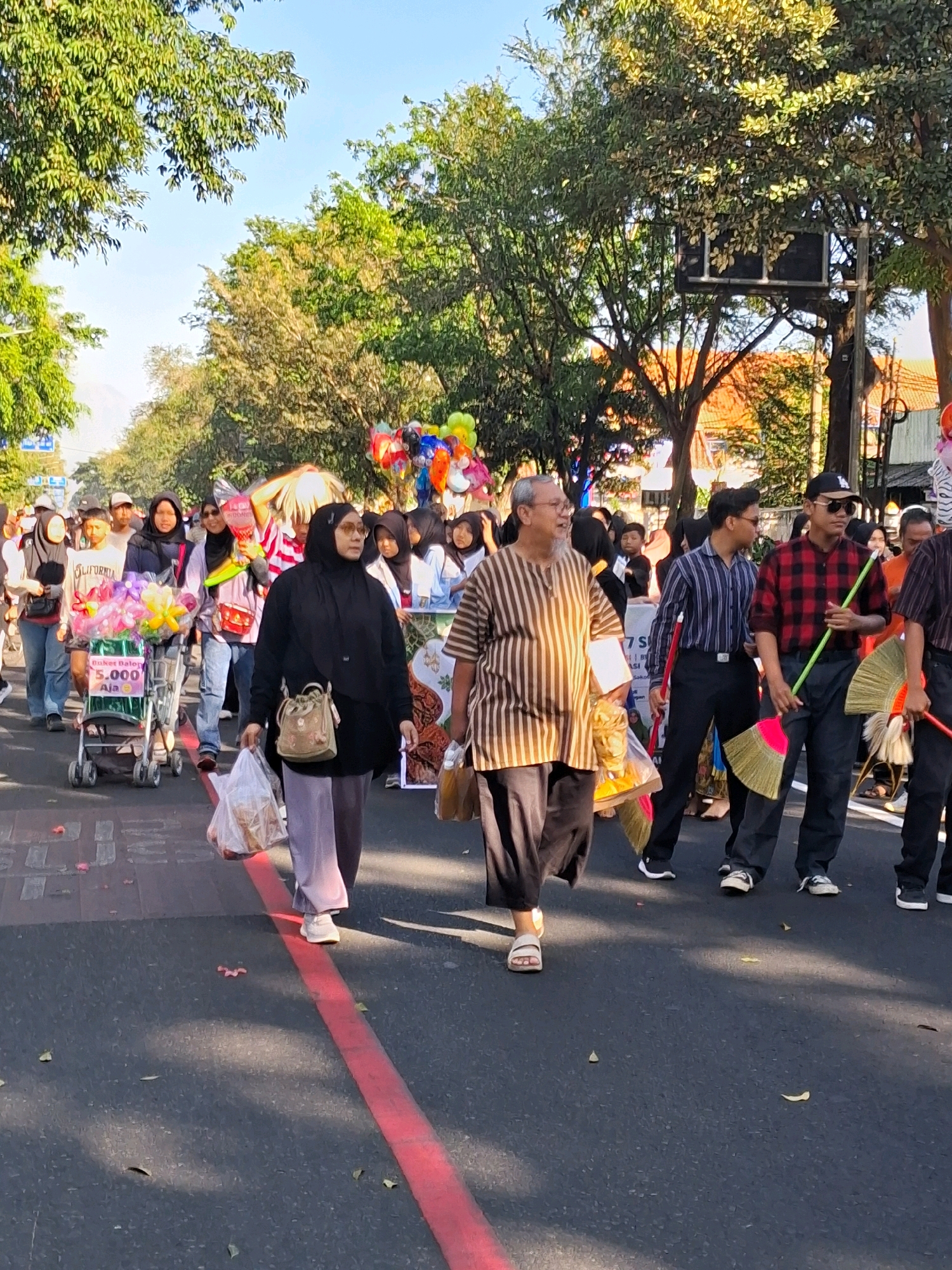 The image depicts a street scene in the city of Surakarta, Indonesia, likely during a parade or festive event. A group of people are walking down a paved street lined with trees on both sides. The sunlight suggests it is daytime.
In the foreground, a woman wearing a black hijab and a long, light-colored skirt walks with a plastic bag in her right hand. She is accompanied by a man in a striped shirt and dark pants, who is also carrying a plastic bag. Behind them, a man in a red and black plaid shirt holds a broom with a green handle, and another man in a striped shirt holds a similar broom.
Several other individuals are visible in the midground and background, including people with hijabs and various clothing styles. There is a shopping cart filled with colorful balloons and flowers, and more balloons are visible further down the street. The presence of balloons and the general atmosphere suggest a celebratory occasion. The street has a red line marking on it. The architecture visible in the background includes tiled roofs.