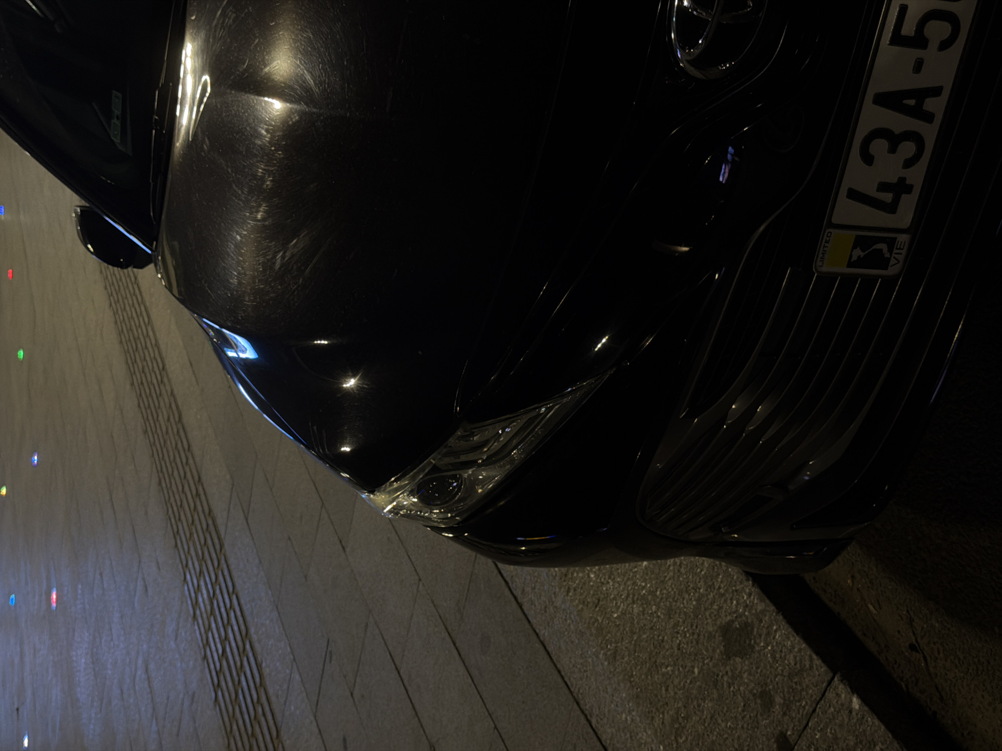 A black car is parked on a paved street at night. The car's front end, including the headlight and grille, is visible, along with a portion of its hood. The license plate, which reads "43A-5", is clearly visible. In the background, colorful lights are embedded in the street, indicating a pedestrian-friendly area or a decorative lighting feature. The pavement consists of square tiles, with a textured strip running horizontally across the image. The scene suggests an urban environment in Da Nang, Vietnam, possibly a commercial district or a public square, given the decorative street lights and tiled pavement. No people are visible in the image.