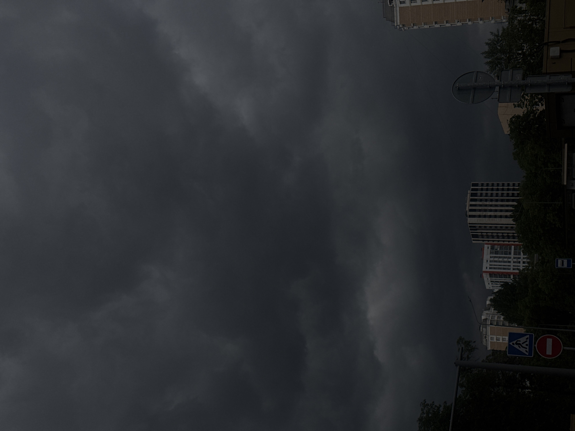 The image depicts a city street in Moscow, Russia, under a dark, overcast sky. Buildings are visible, including high-rise residential structures with multiple floors and some older, likely Soviet-era apartments. Street signs are present, including pedestrian crossing and no-entry signs. Trees and other vegetation line the street. The dark sky and dim lighting suggest a storm or approaching nightfall. No people are visible in the image.