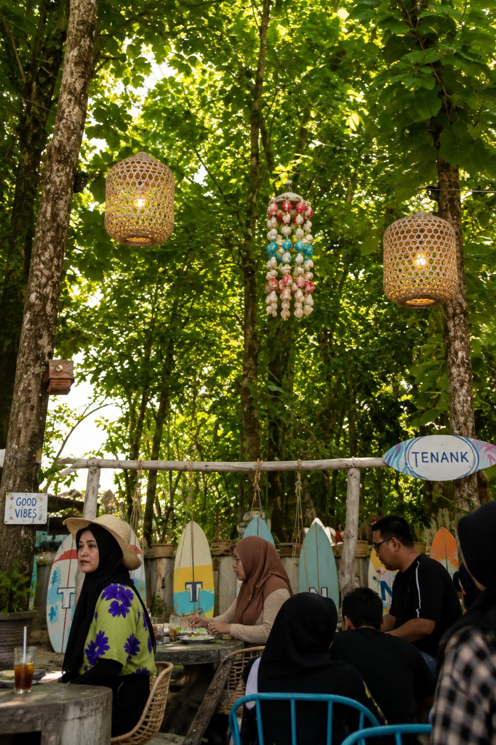 The image captures an outdoor seating area under a canopy of lush green trees, likely in the city of Medan, Indonesia, given the context of the "TENANK" sign which is a popular cafe in Medan. The scene is set at a cafe with a relaxed, tropical ambiance.
In the foreground, a woman wearing a black hijab and a straw hat is seated at a table, looking to her left. She is dressed in a vibrant lime green shirt with purple floral patterns. On the table in front of her is a glass with a dark beverage and a straw. To her right, a woman in a light brown hijab is seated at another table, engaged in eating and looking down at her food. She is wearing a beige cardigan. Behind them, other patrons are visible, including a man with glasses in a black t-shirt and several individuals partially obscured, one of whom is wearing a black headscarf.
The environment is decorated with surfboards arranged vertically behind the seating area, adding to the beach-themed decor. Two woven pendant lights hang from the trees, one on the left and another on the right, casting a warm glow. A colorful beaded mobile, featuring red, white, and blue beads, hangs between them. A sign with the word "TENANK" in a wavy, light blue font is affixed to a wooden pole. Another smaller sign on a tree trunk to the left reads "GOOD VIBES." The overall atmosphere suggests a casual dining or gathering spot amidst nature.