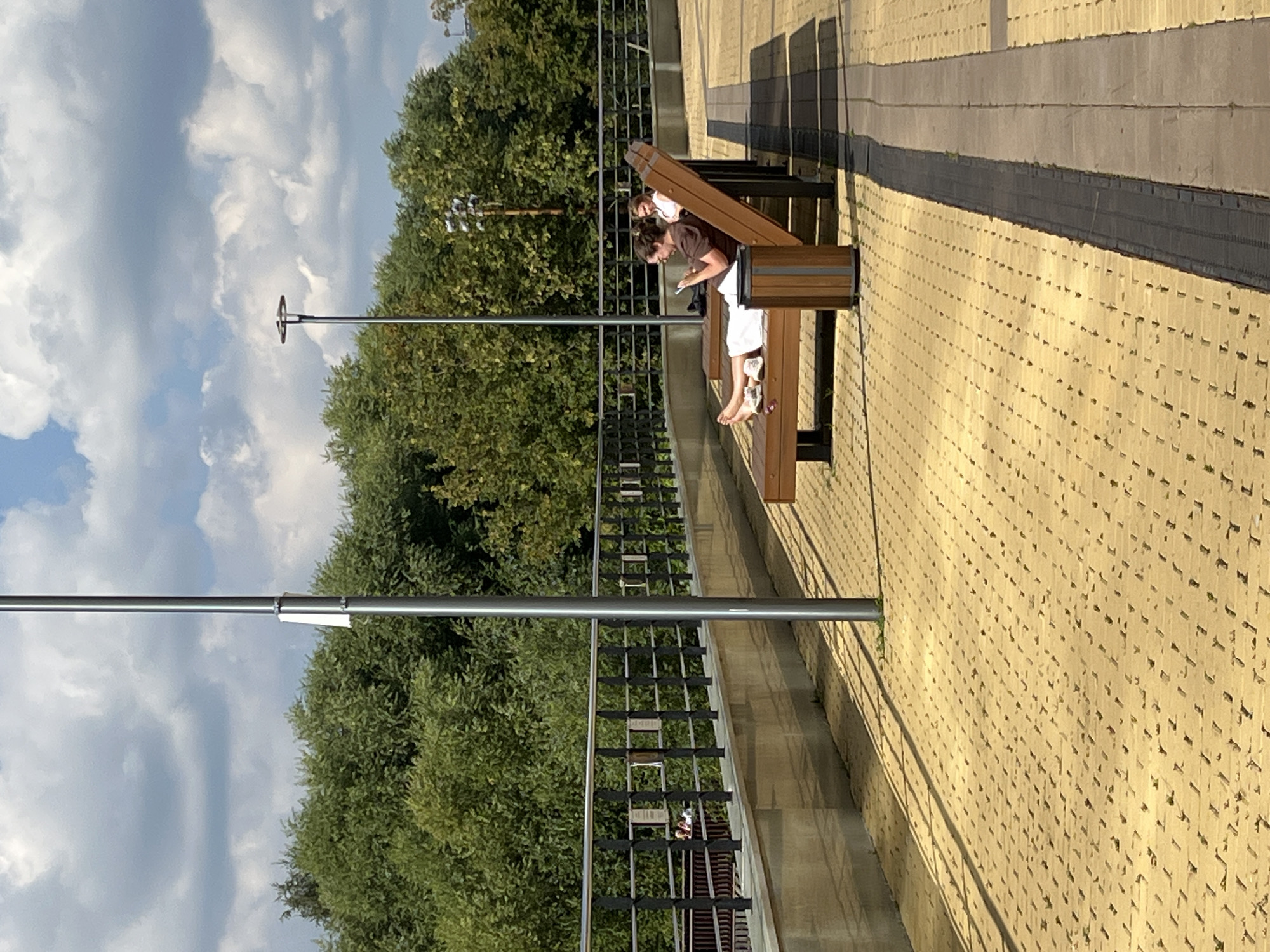 The image shows a paved public space with benches and trees. A woman is sitting on a bench, looking at her phone. She is wearing a brown shirt and white shorts, and her feet are bare. There is another person sitting on a bench in the background. The space appears to be a park or promenade, with a railing and a wall in the background. The sky is cloudy, and the sun is casting shadows on the ground.
This image was taken in Yekaterinburg, Russia. Specifically, it is a view of the embankment along the Iset River, a popular spot for recreation and relaxation in the city. The paved area is likely part of a newly developed or renovated section of the embankment, featuring modern benches and lighting. The trees in the background are characteristic of the vegetation found along the river. The railing is likely a safety feature for the embankment, and the wall might be part of a retaining structure or a raised platform.