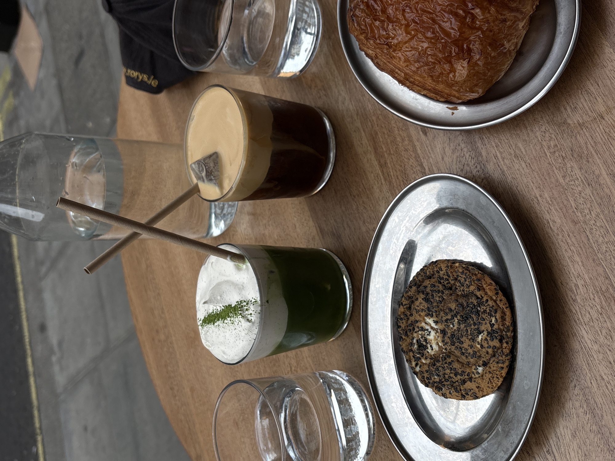 A top-down shot displays a wooden table with a variety of food and beverages, possibly in London, United Kingdom, given the website "storys.ie" on a cap. On the table are two stainless steel plates; one holds a flaky, brown pastry, likely a croissant, and the other contains a round, textured cookie sprinkled with black sesame seeds. There are two glasses of drinks: one with a dark bottom and a cream-colored top, containing a straw, and another with a green liquid at the bottom and a white, frothy top garnished with green powder, also with a straw. A half-filled glass water bottle lies horizontally, while two additional empty glasses complete the setting. Part of a black cap with white lettering can be seen at the top of the image, near the edge of the table. The background shows a glimpse of a grey sidewalk.