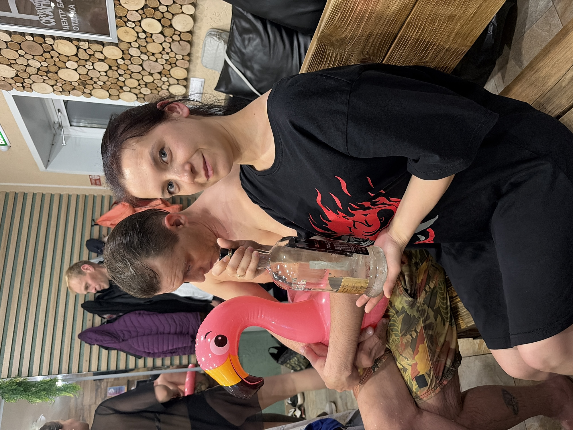 Here is a detailed description of the image:
**Scene:** The image appears to be taken indoors, possibly in a sauna or relaxation area. The setting is characterized by light-colored tiled floors and walls lined with wooden planks, some arranged decoratively with circular wood pieces.
**People:** Two individuals are prominent. A woman with dark hair is seated, facing the camera, wearing a black t-shirt with a red flame design and black shorts. A man is also present, holding a half-empty glass bottle and an inflatable pink flamingo. At least two more people are visible in the background, one standing and another possibly seated.
**Objects:** A key object is the half-empty glass bottle of liquid held by the man, as well as the inflatable pink flamingo he is holding. There is a wooden bench or seating area near the woman. Other visible objects include clothing items in the background (a purple jacket, a black sheer garment), and a wall-mounted sign with text in Russian, partially visible in the upper left corner.
**Actions/Interactions:** The man appears to be pouring from the bottle into the flamingo, suggesting a playful or humorous action. The woman is looking directly at the camera, possibly posing for the picture.
**Environment:** The presence of wooden benches, wood-paneled walls, and tiled floors suggests a sauna or similar relaxation space. The decor is simple and functional, with some decorative elements like the wooden circular accents on the wall.
