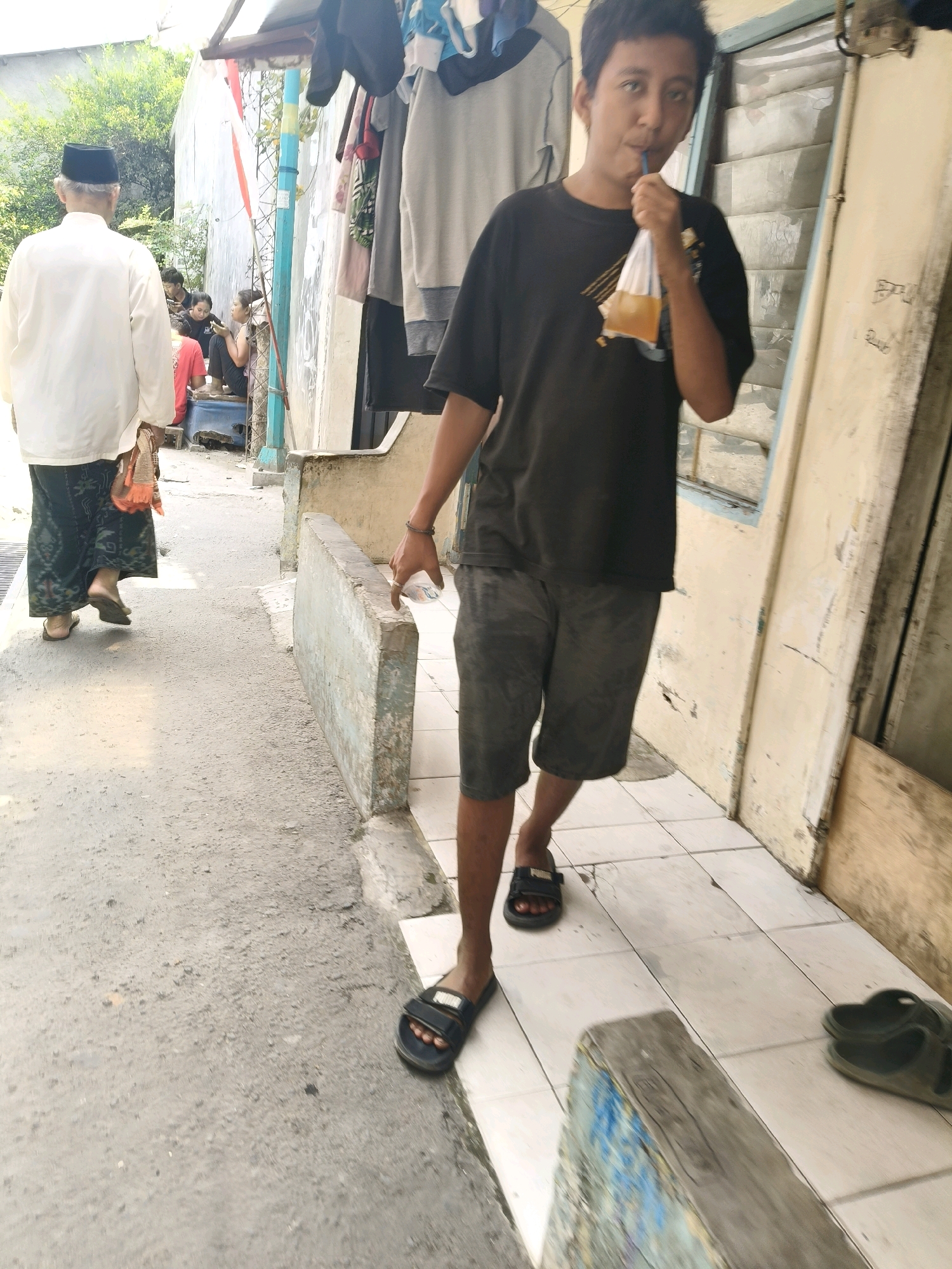 In an outdoor alleyway in South Tangerang, Indonesia, a young man in a dark t-shirt and grey shorts is drinking from a plastic bag with a straw. He is walking on a tiled walkway next to a concrete wall. Further down the alley, an older man wearing a white shirt and black sarong walks away from the camera, carrying a bag. Other people are visible in the background, seated near a building. Clothes are hanging on a line overhead.
