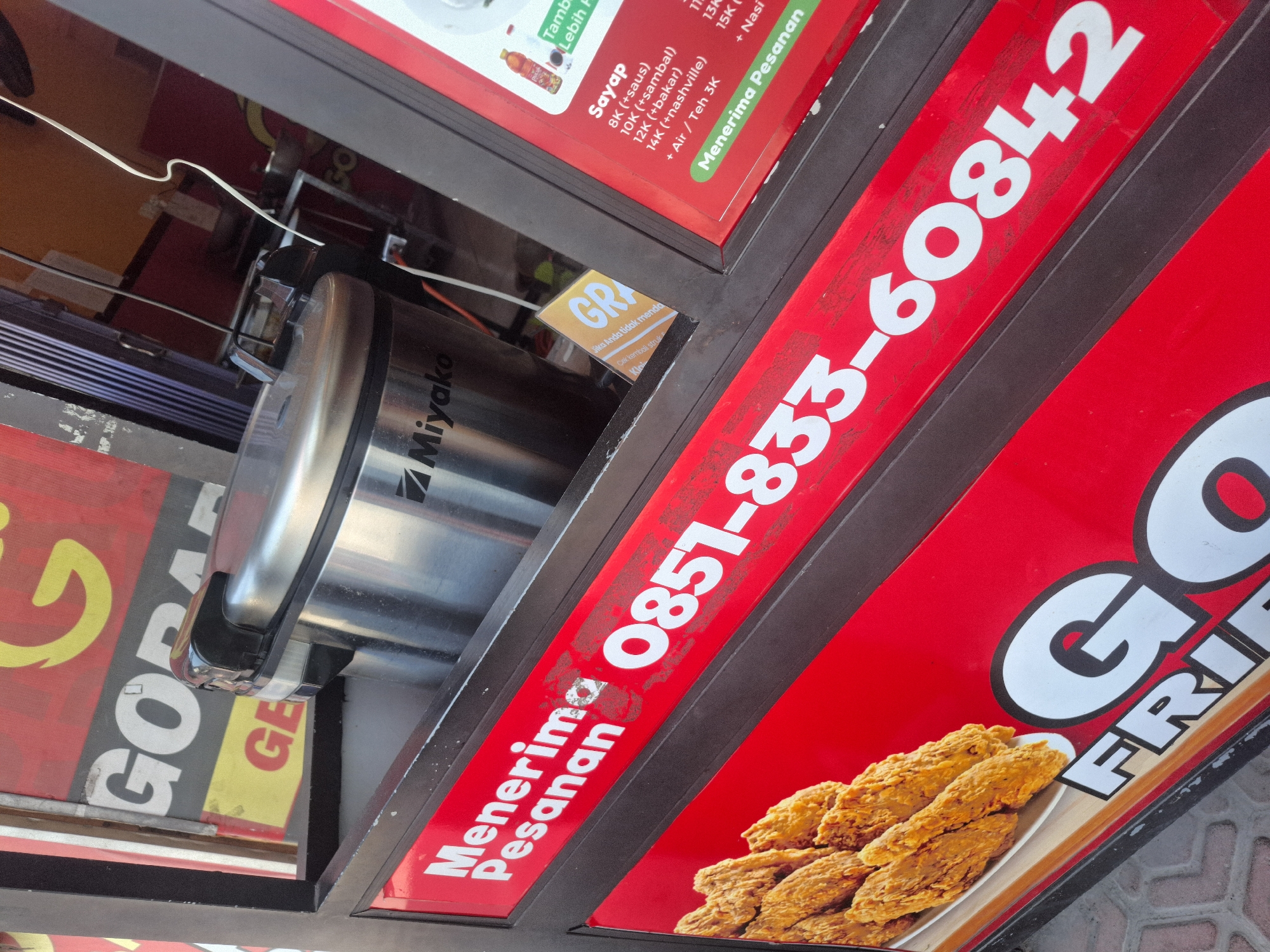The image shows a close-up of a food stall in Sumberwudi, Indonesia. The stall appears to be selling fried chicken, as evidenced by a picture of fried chicken wings on the left side of the image. Prominently displayed on a red banner are the words "Menerima Pesanan" (Accepting Orders) and a phone number, "0851-833-60842". Above this, a silver rice cooker with the brand "Miyako" is visible. A menu card with prices for "Sayap" (Wings) and various sauces is also visible on the right. The background suggests an indoor setting with a dark shuttered door and some signage. The overall impression is a functional, roadside food establishment.