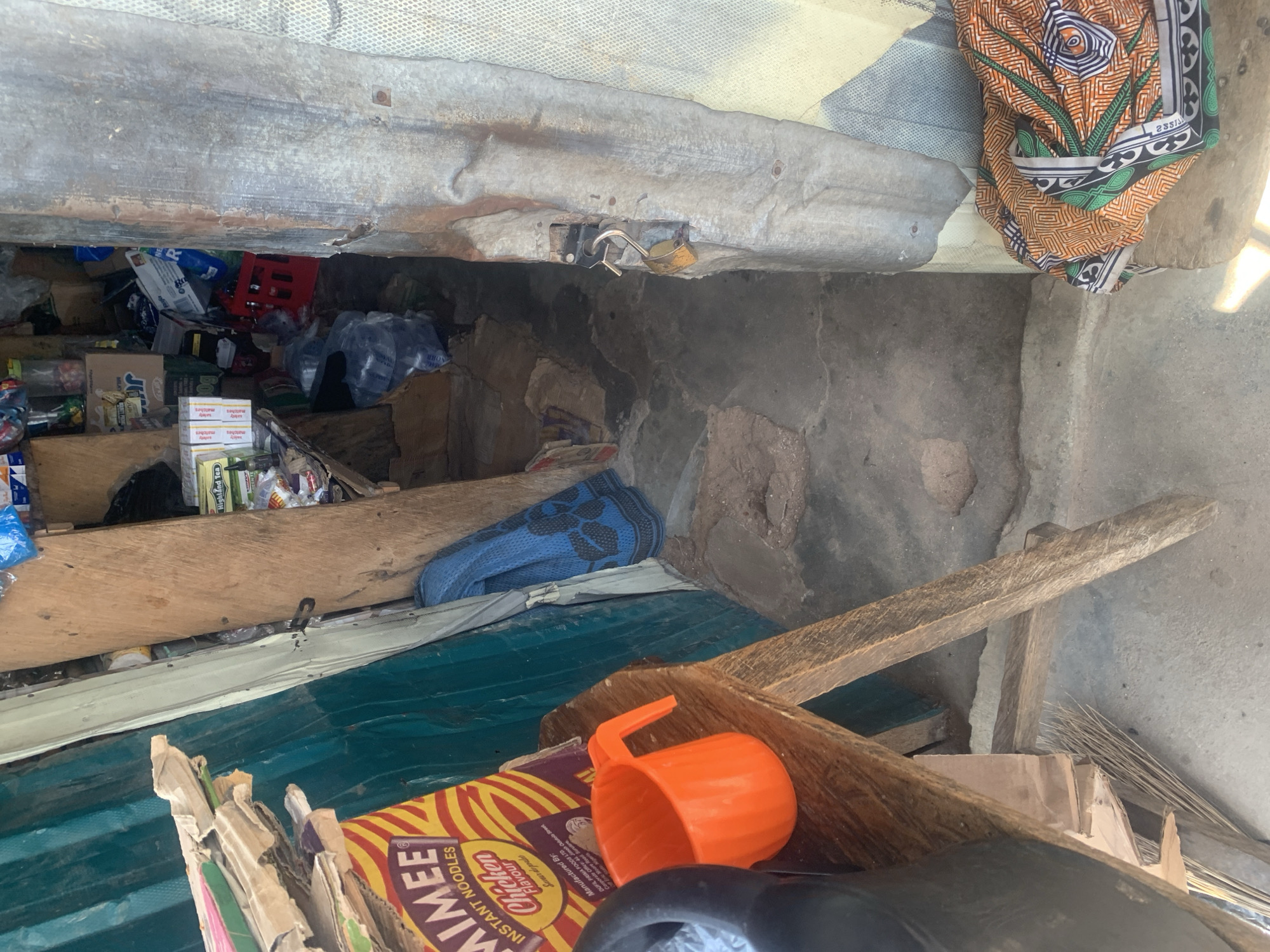 The image shows a cluttered outdoor stall or shop in Jalingo, Nigeria. The stall appears to be made of corrugated metal and wood, with various items stacked inside. In the foreground, there are boxes of instant noodles labeled "MIMEE Chicken Flavour" and an orange plastic mug. Behind these items, a blue cloth is visible, and further back, shelves are filled with packaged goods, including what looks like tea boxes and other food items. A red crate is also visible among the inventory. The overall impression is of a small, informal business operation. The lighting suggests it is daytime. There is no clear indication of any specific event or activity, beyond the general appearance of a stocked stall. The scene is characterized by a sense of disorganization and the use of basic materials for construction.