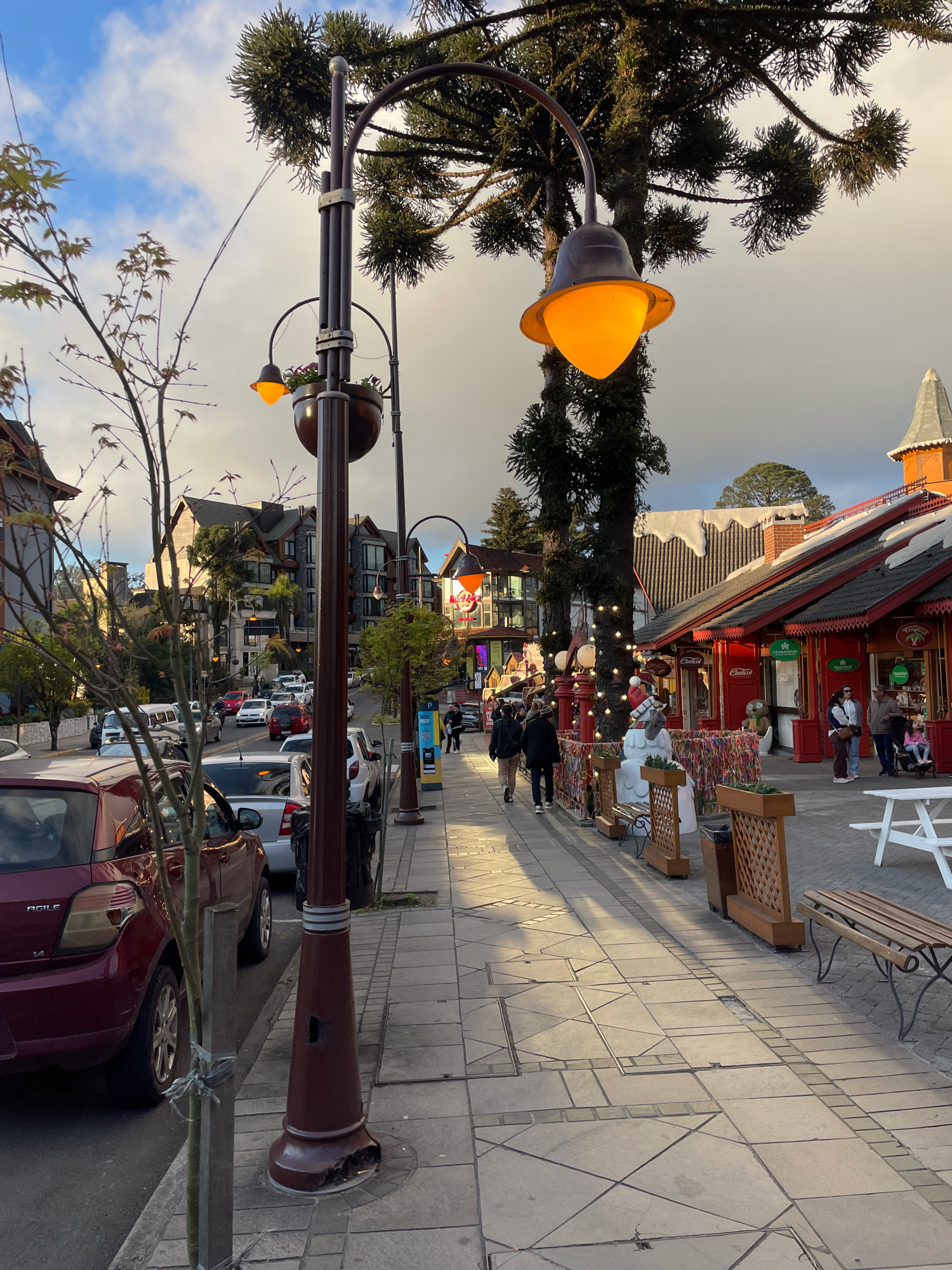 The image depicts a street in Gramado, Brazil, lined with shops and buildings on the right, and parked cars and a sidewalk on the left. A prominent lamppost with an orange light stands in the foreground. Several people are walking on the sidewalk. The street appears to be paved with cobblestones and tiles. The buildings on the right have red roofs and facades decorated with Christmas lights, suggesting a festive atmosphere. Trees, including a large conifer, are visible. The sky is cloudy with patches of sunlight.