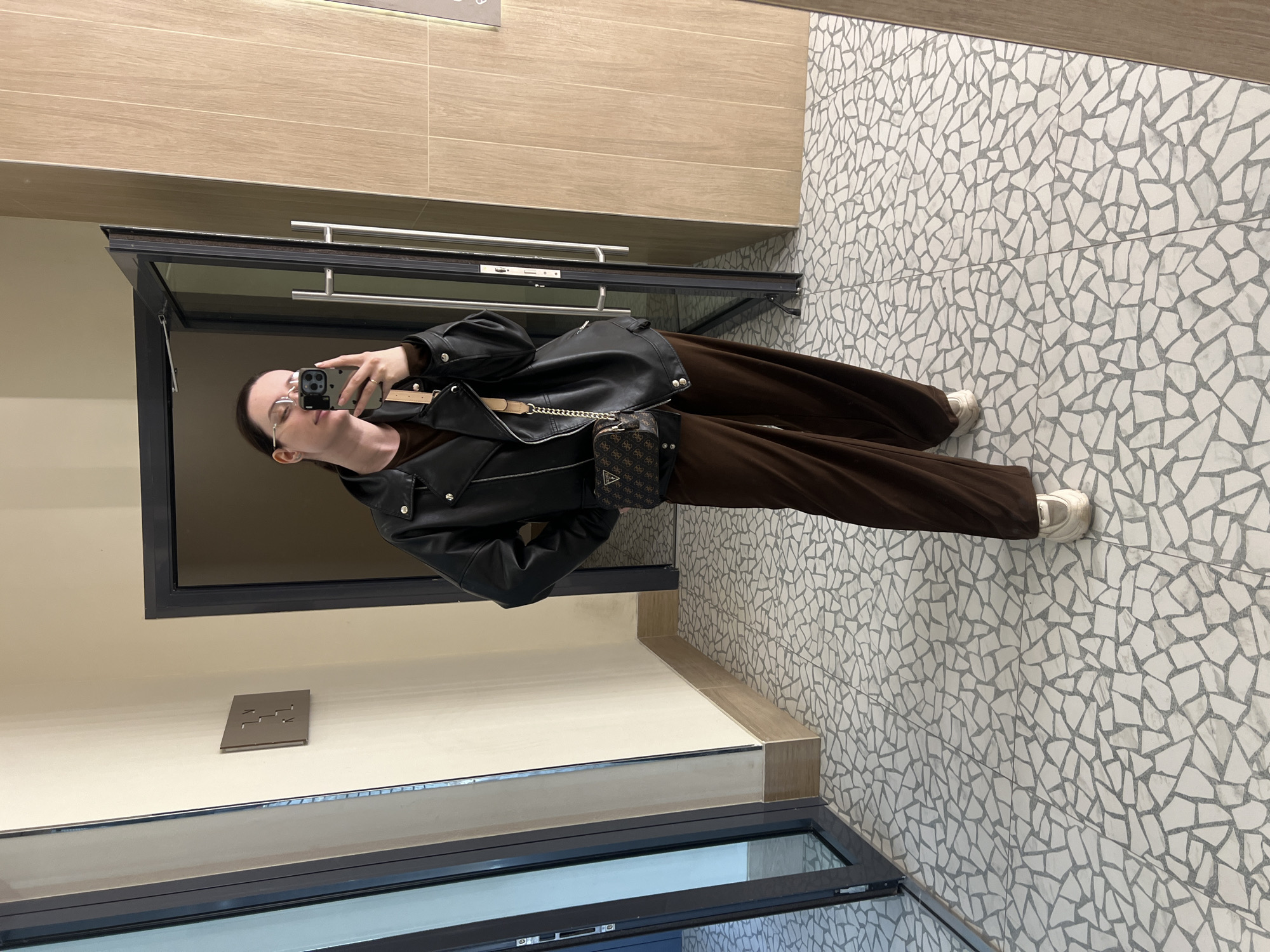 The image shows a woman taking a picture of herself in a mirror. She is wearing glasses, a black leather jacket, brown pants, and white sneakers. She is also carrying a small purse with a chain strap. The background is a bathroom with beige wall tiles and mosaic floor tiles. The location is Tyumen, Russia. A metal grate is visible in the wall underneath the mirror.