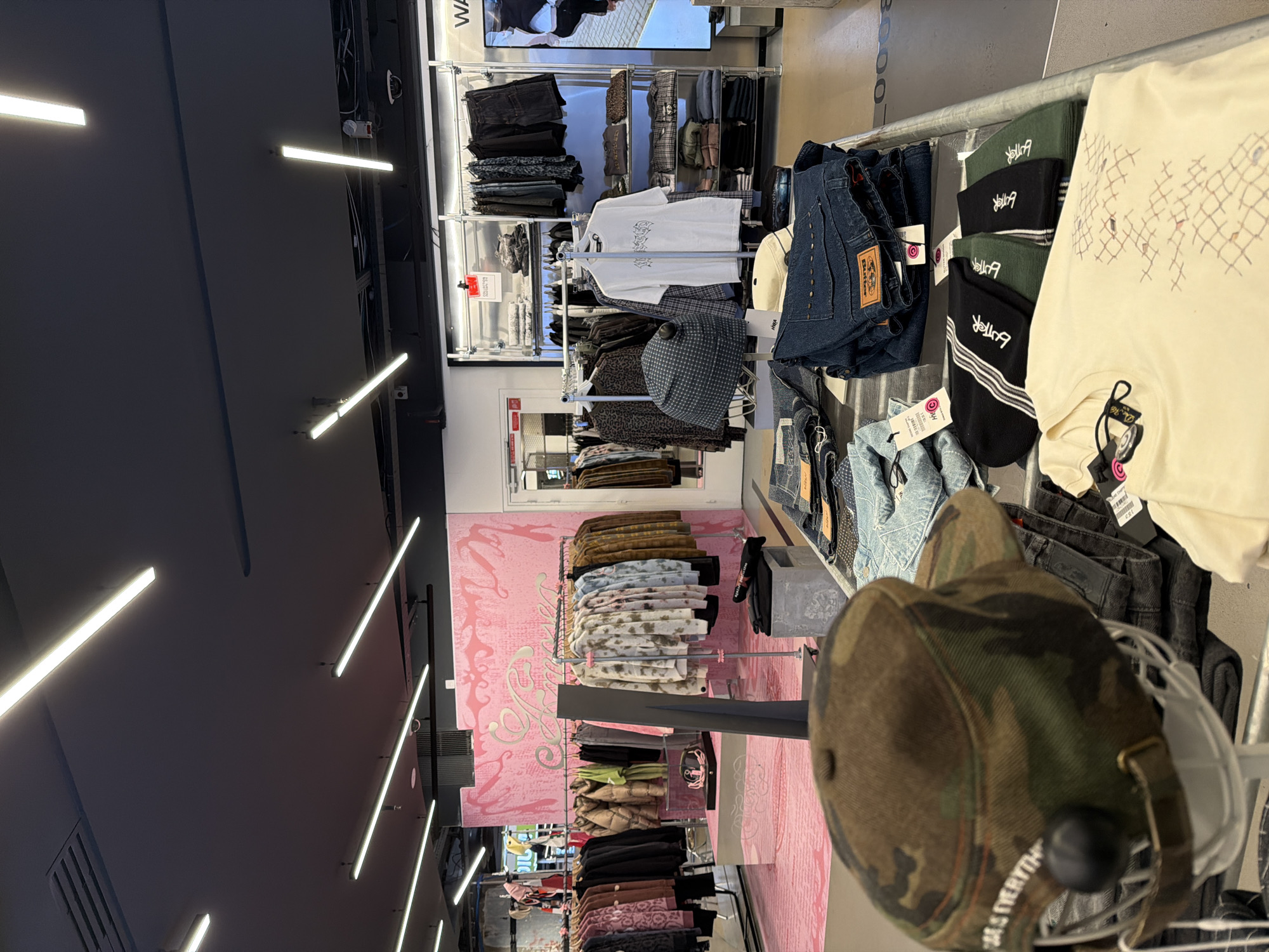 The image depicts the interior of a clothing store in Paris, France. The store is well-lit with fluorescent ceiling lights. Clothing is displayed on racks along both sides of the aisle. There are shirts, jeans, and other garments on display. A pink wall with a decorative pattern is visible on one side. In the foreground, a camouflage baseball cap is prominently displayed on a rack.