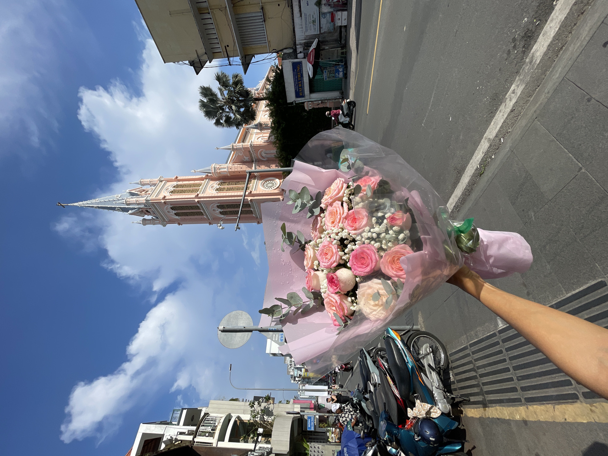 The image displays a sunny urban street scene in Ho Chi Minh City, Vietnam. In the foreground, a tan-skinned forearm extends from the lower right, holding a large bouquet of flowers. The bouquet contains approximately 15-20 pink and peach roses, interspersed with white baby's breath and green eucalyptus leaves, all wrapped in a clear plastic film over light pink paper, with small water droplets visible on the plastic. The arm casts a shadow onto the dark gray tiled sidewalk, which features a long drainage grate.
The midground reveals a bustling city street. On the left, multiple motorbikes of varying models and colors are parked diagonally along the sidewalk. Adjacent to these are multi-story buildings with balconies, one bearing a blue street sign that reads "ĐƯỜNG DINH CÔNG TRÁNG". A lamppost and a round gray traffic sign on a pole stand near the center-left. The street itself is asphalt with a double yellow line running down its center. On the right side of the road, a person on a white scooter rides away from the viewer.
The background is dominated by the distinctive pink Gothic-style Tân Định Church (Church of the Sacred Heart of Jesus), which features a tall, slender steeple topped with a cross and multiple turrets. A large palm tree stands to the right of the church structure, partially obscuring a two-story yellow building. A sign above a gate near the church reads "TRƯỜNG PHỔ THÔNG HUYỀN HAI BÀ TRƯNG". The sky is bright blue with scattered white cumulus clouds.