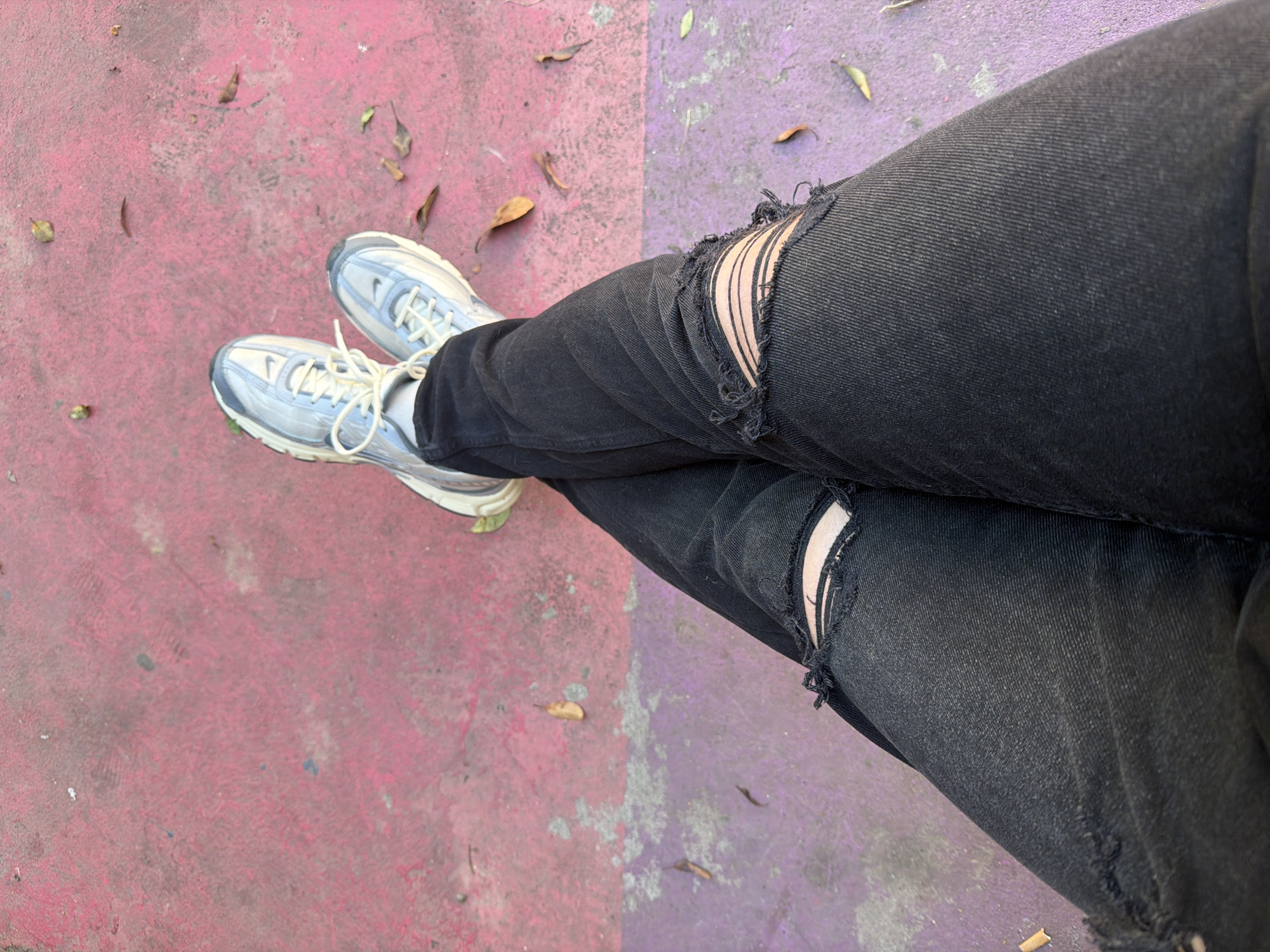 A person wearing ripped black jeans and white sneakers is sitting on a surface that is divided into two colors, purple and red. The person's legs are crossed at the knee. There are some leaves scattered on the surface. The image provides no information regarding the specific location within the Palilula Urban Municipality or any additional details about the individuals involved.