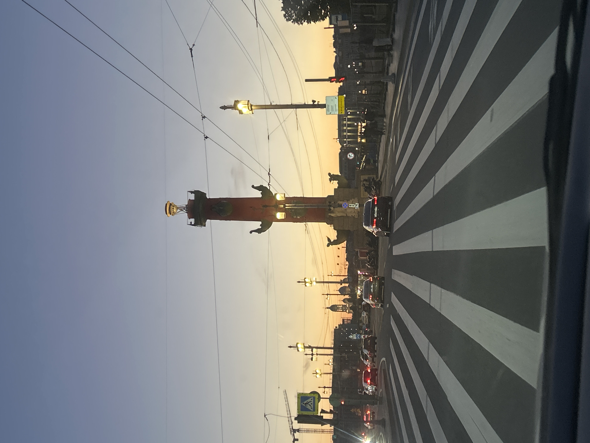A wide-angle, low-angle shot captures a street scene at dusk in an urban environment, possibly St. Petersburg, Russia. The sky transitions from a pale blue at the top to a warm orange and yellow gradient near the horizon. A prominent Rostral Column, a tall, reddish-orange structure topped with a lantern, stands in the middle ground. Several streetlights with illuminated globes line the street, casting a warm glow.
A busy intersection features a pedestrian crossing with bold white stripes painted across the dark asphalt. Cars of various colors, including black, silver, and red, are moving through the intersection, some of them appearing to be in traffic. A pedestrian crossing sign with a blue background and a white silhouette of a person is visible on the left. In the distance, a crane is visible, suggesting ongoing construction. The overall impression is that of a lively city street during the evening rush hour.