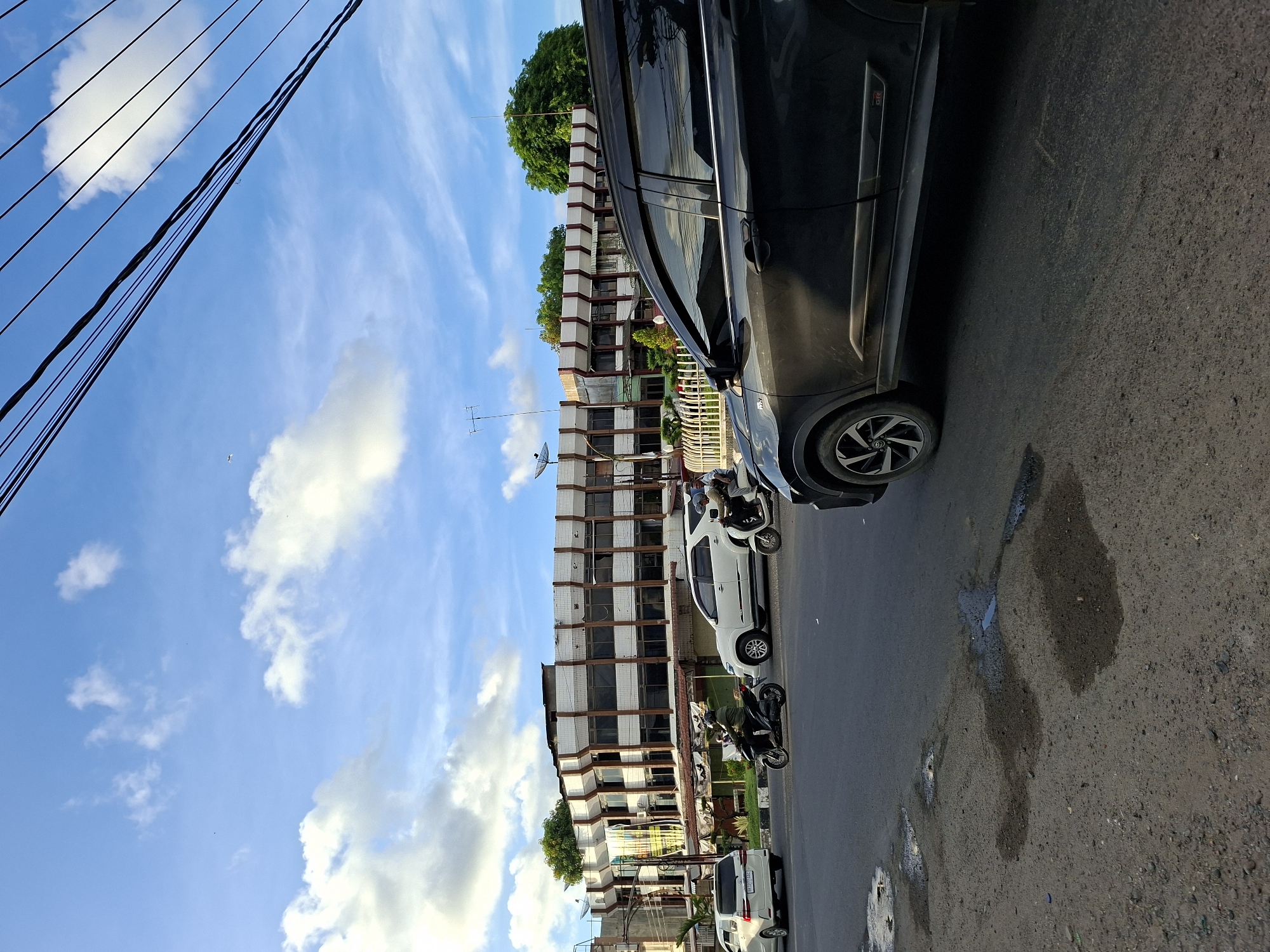 The image depicts a street scene in Lhokseumawe, Indonesia. The foreground is dominated by a black car parked on the right side of the street. Several motorcycles and other vehicles are visible parked along the side of the road. In the background, a multi-story building with a white facade and brown trim is prominent. Utility lines are strung across the sky, which is partly cloudy with a blue hue. The street pavement shows signs of wear and possibly recent water accumulation.