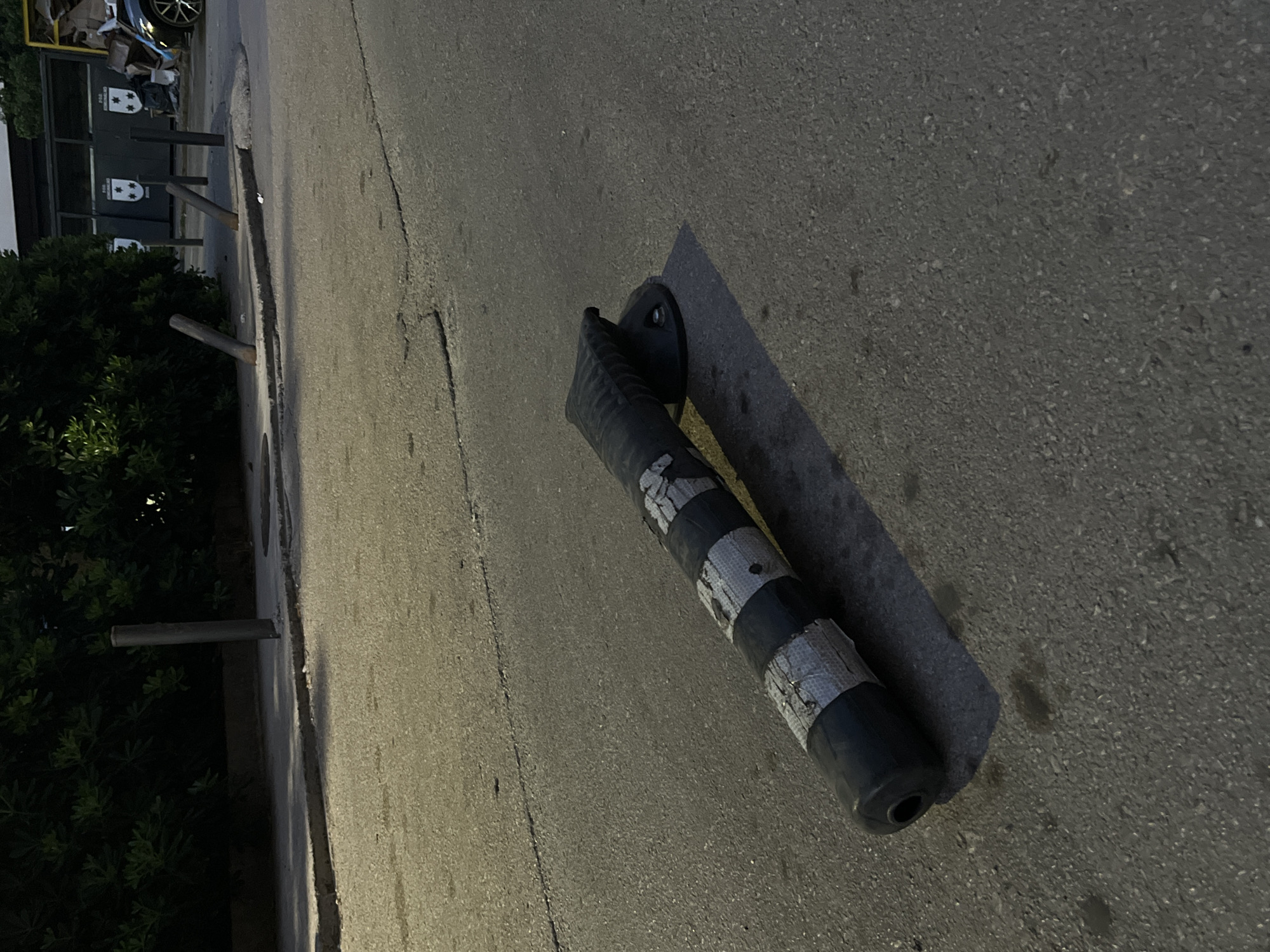 The image shows a damaged, black and white striped traffic bollard lying on its side on an asphalt road. The bollard appears to be made of plastic and is broken or bent near its top. Silver reflective tape is wrapped around the bollard in several sections. A black circular base is attached to the wider end of the bollard. In the background, there is a sidewalk with several metal posts, some of which are leaning. Behind the sidewalk is a dense hedge of dark green foliage. Further in the background, there are large, dark-colored recycling bins and a portion of a building. A black car is visible to the far right. The scene is dimly lit, suggesting it is either dusk or early morning. The location is identified as Budva, Montenegro.