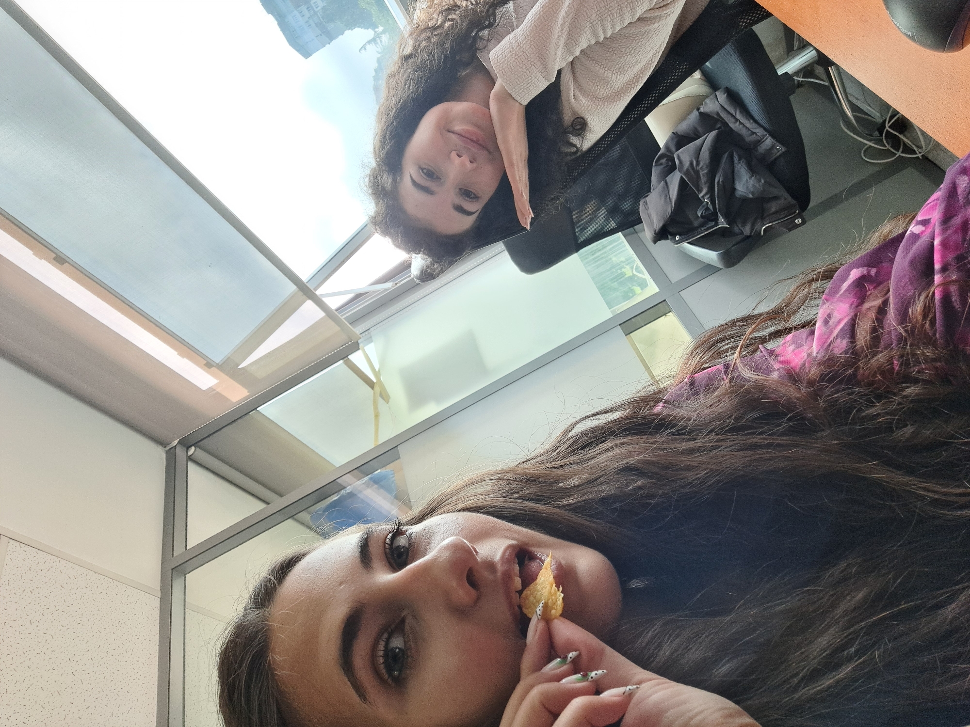 This image, taken in Sofia, Bulgaria, captures two young women within what appears to be a modern office or co-working space. The photo is rotated sideways and taken from a low perspective, looking upwards.
In the foreground, a woman with long, dark, wavy hair and light eyes is prominently featured. She is in the process of eating a potato chip, holding it to her mouth with a hand showcasing manicured nails with a white and black dotted design. She wears a top with a visible purple and pink pattern.
Further back, another woman with curly brown hair and light eyes is seated, leaning on her arm. She looks towards the camera with a gentle, relaxed smile. She is dressed in a light-colored, textured long-sleeved top. Behind her, a black office chair is visible, and a black puffer jacket is draped over another chair. A light-colored desk surface is partially seen, along with a black computer mouse and tangled cables on the floor.
The setting is characterized by large windows that reveal a bright daytime sky and the architectural facade of other buildings, indicative of an urban environment. The interior features modern glass partitions with grey frames and white, textured ceiling tiles. The overall scene suggests a casual moment or a break from work during the day. No text is visible in the image.
