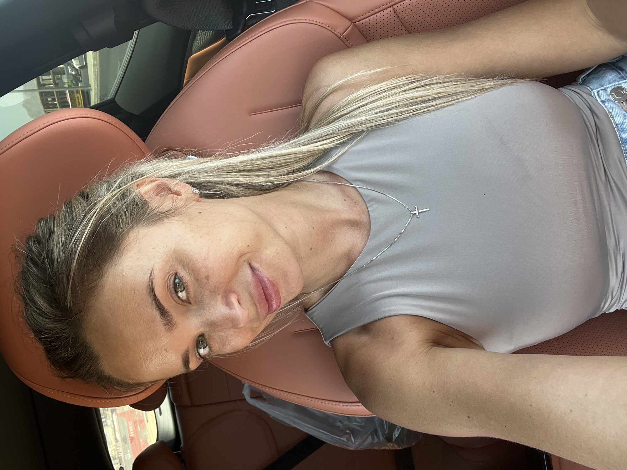 A Caucasian woman with blonde hair pulled back, freckles, and light green eyes is in the driver's seat of a car. She is looking at the camera with a slight smile. She is wearing a grey sleeveless top and a silver necklace with a cross pendant. The interior of the car is visible, with brown leather seats. Outside the car window, a street scene in Sharm El Sheikh, Egypt, is depicted, with buildings and vehicles visible.