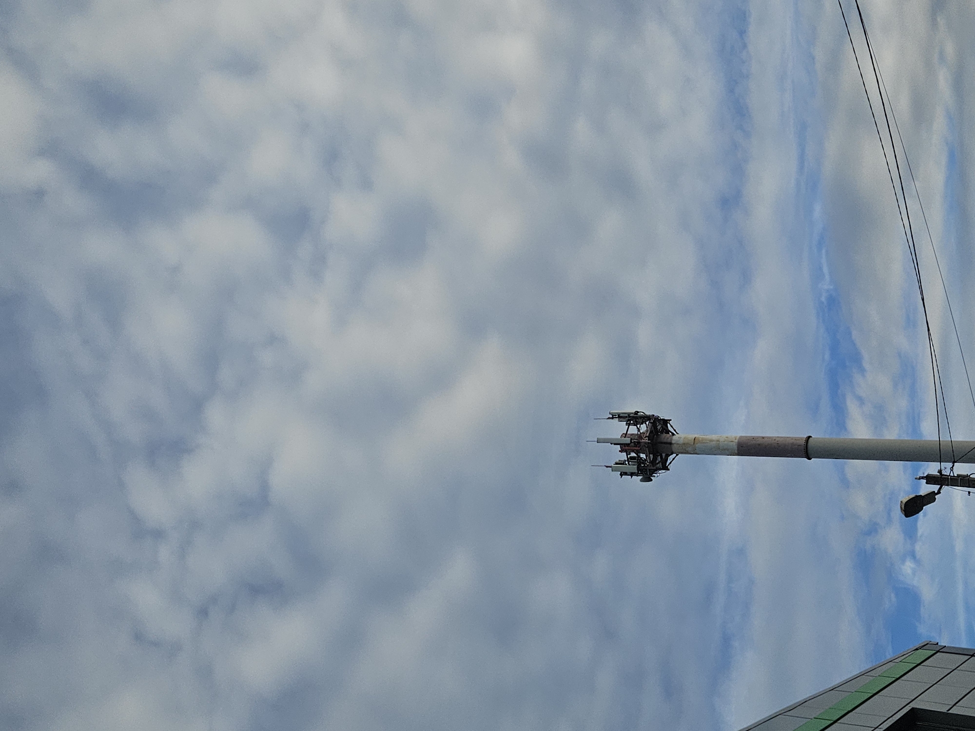 The image shows a cell tower with antennas against a backdrop of a cloudy sky. Part of a building with a gray roof and green accents is visible in the lower right corner. Power lines run down the right side of the frame. The tower appears to be in Осеченки, Russia.