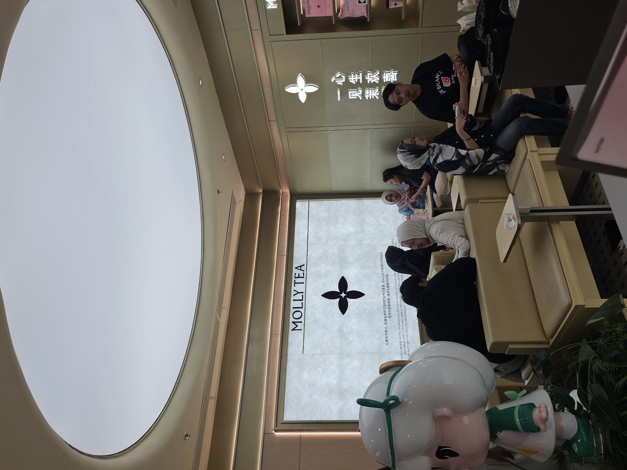 In a brightly lit interior space, likely a retail establishment, several individuals are seated on plush, cream-colored benches, suggesting they are waiting. The scene is dominated by a large, translucent window or display panel featuring the text "MOLLY TEA" in bold, black lettering, accompanied by a stylized four-petal flower logo. Above this, Chinese characters are prominently displayed, which translate to "happiness in the heart" and "meet jasmine."
A young man in a black t-shirt stands near the display, looking towards the camera. A woman in a patterned headscarf sits on a bench, also looking away from the camera. Other people are visible on the benches, some wearing headscarves, indicating a potentially cultural gathering or a pause in their shopping experience.
The ceiling features a large, circular, illuminated dome, casting a soft, diffused light on the environment. The overall atmosphere appears calm and relaxed. The visible text "MOLLY TEA" suggests the establishment is a tea shop or a brand associated with tea. The style of the decor and the presence of people waiting imply a modern commercial setting. The bright lighting and clean aesthetic point towards a daytime scene, possibly late afternoon given the general ambiance. The presence of plants in the foreground adds a touch of greenery to the scene, further enhancing the welcoming atmosphere.