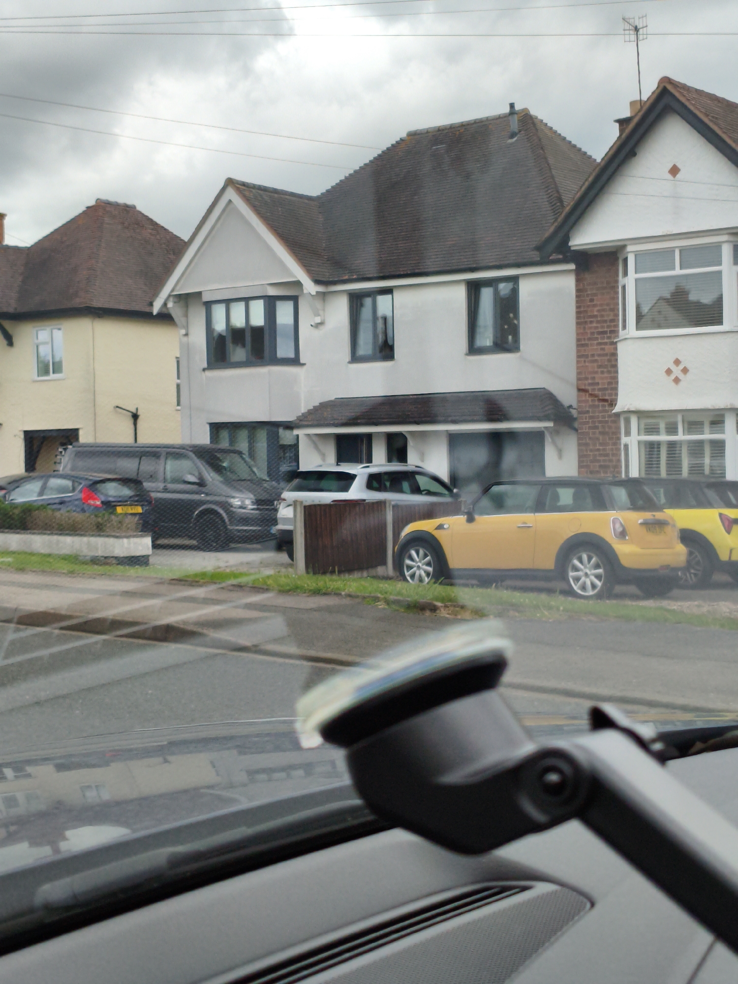 A daytime, medium-shot, exterior view shows a residential street scene in Wychavon, United Kingdom. The focus is on several parked cars, including a black van, a yellow Mini Cooper, and a silver SUV, positioned in front of residential properties with overcast skies above. The foreground of the image is dominated by the interior of a vehicle, with a dashboard and windshield wipers visible, suggesting the photo was taken from a car. The scene conveys a sense of ordinary urban life.