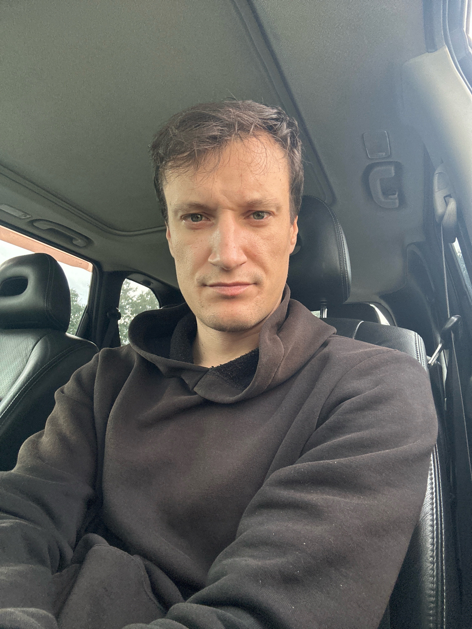 A man with short, dark hair and light skin is sitting in the driver's seat of a car. He is wearing a dark brown hooded sweatshirt. His arms are crossed, and he is looking directly at the camera with a neutral expression. The interior of the car is visible, including the black leather seats, the sunroof, and the headliner. The background outside the car shows trees. The image was taken outdoors in the city of Makiv, Ukraine.