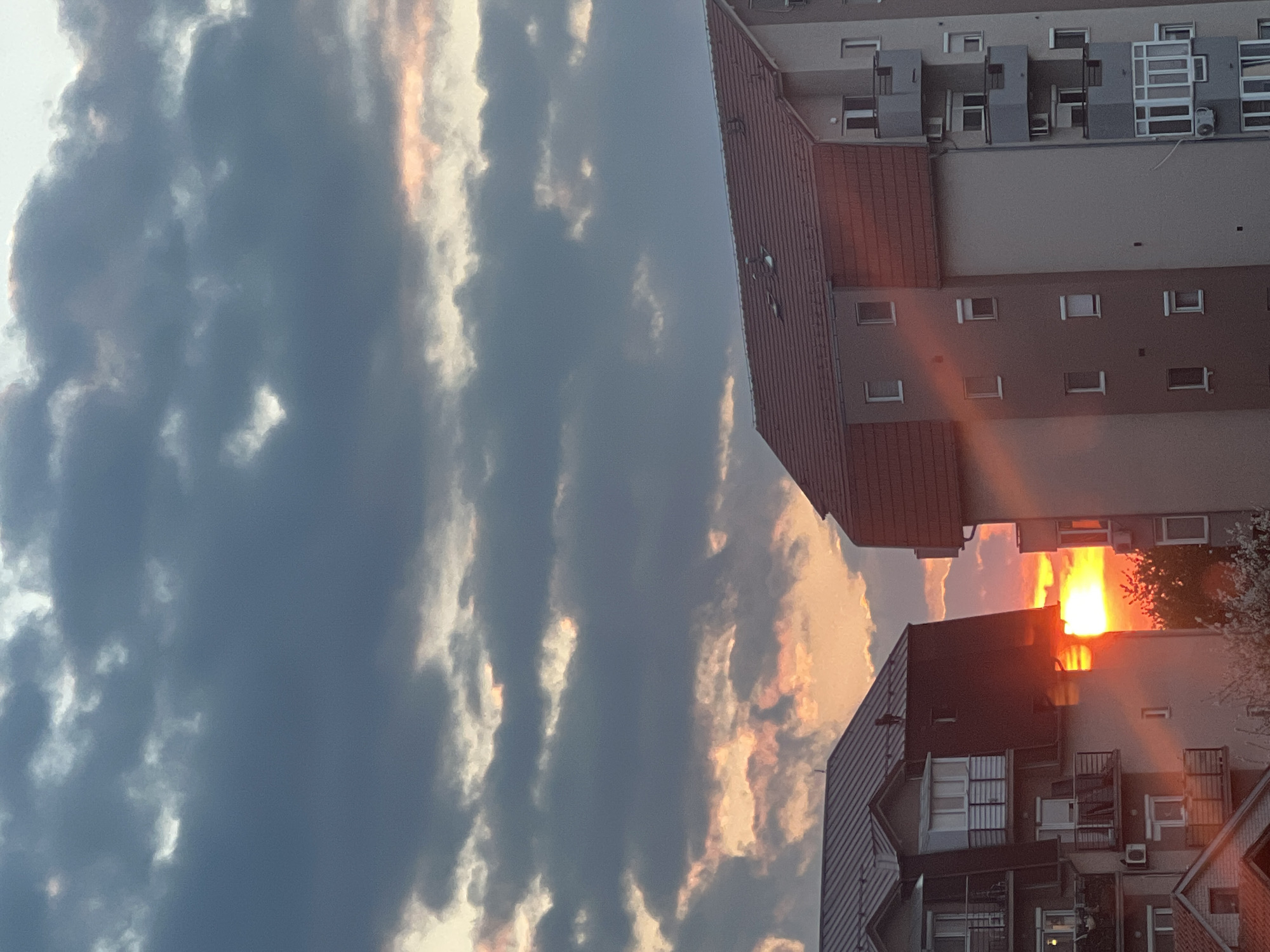 The image depicts a sunset in Palilula Urban Municipality, Serbia. The sky is filled with cumulonimbus clouds, some illuminated with orange light from the setting sun. Adjacent to the sky are several buildings. One is a multi-story apartment building with a red tiled roof and multiple rectangular windows, another is a building lower to the ground, also with a red tiled roof. A ray of direct sunlight shines on the taller building.