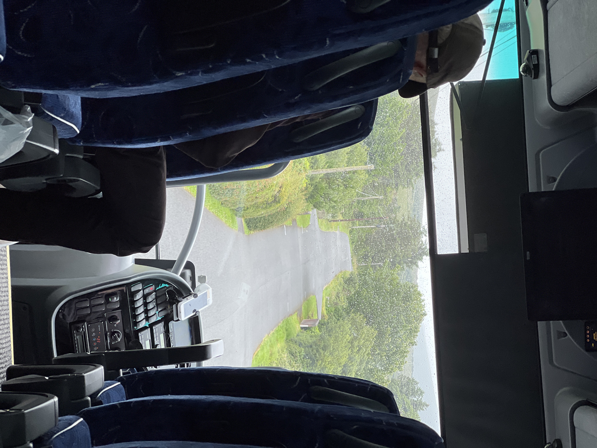 The image provides an interior view from a bus, looking forward through the windshield. The road ahead is wet and appears to be a rural, winding path surrounded by lush green trees and vegetation, with a gray sky suggesting overcast or rainy weather. In the foreground, the dark blue seats of the bus are visible, and a person wearing a baseball cap is seated on the left. The bus's dashboard, with various buttons and a mounted phone, is also visible. The scene is captured from within the bus, implying a journey is in progress. The location is identified as Cearnóg Sheain Bháin, Ireland.