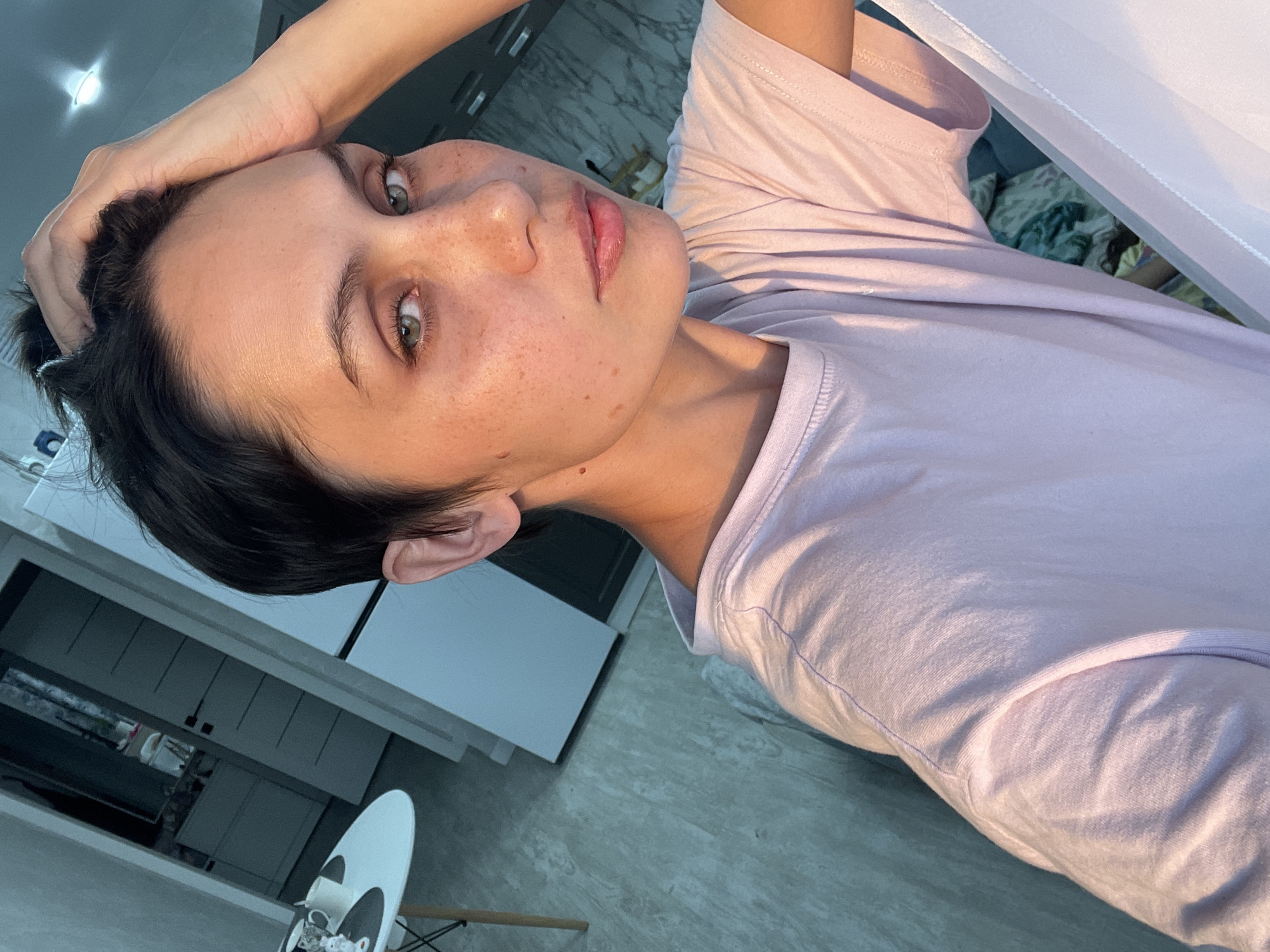 A close-up portrait shows a fair-skinned person with dark hair. The person's hand is on their head. Freckles are visible on the face. The person wears a light pink t-shirt. The background includes a light-colored wall, kitchen cabinets, a marble counter, and a table. The location is not identifiable as being in Izhevsk, Russia.