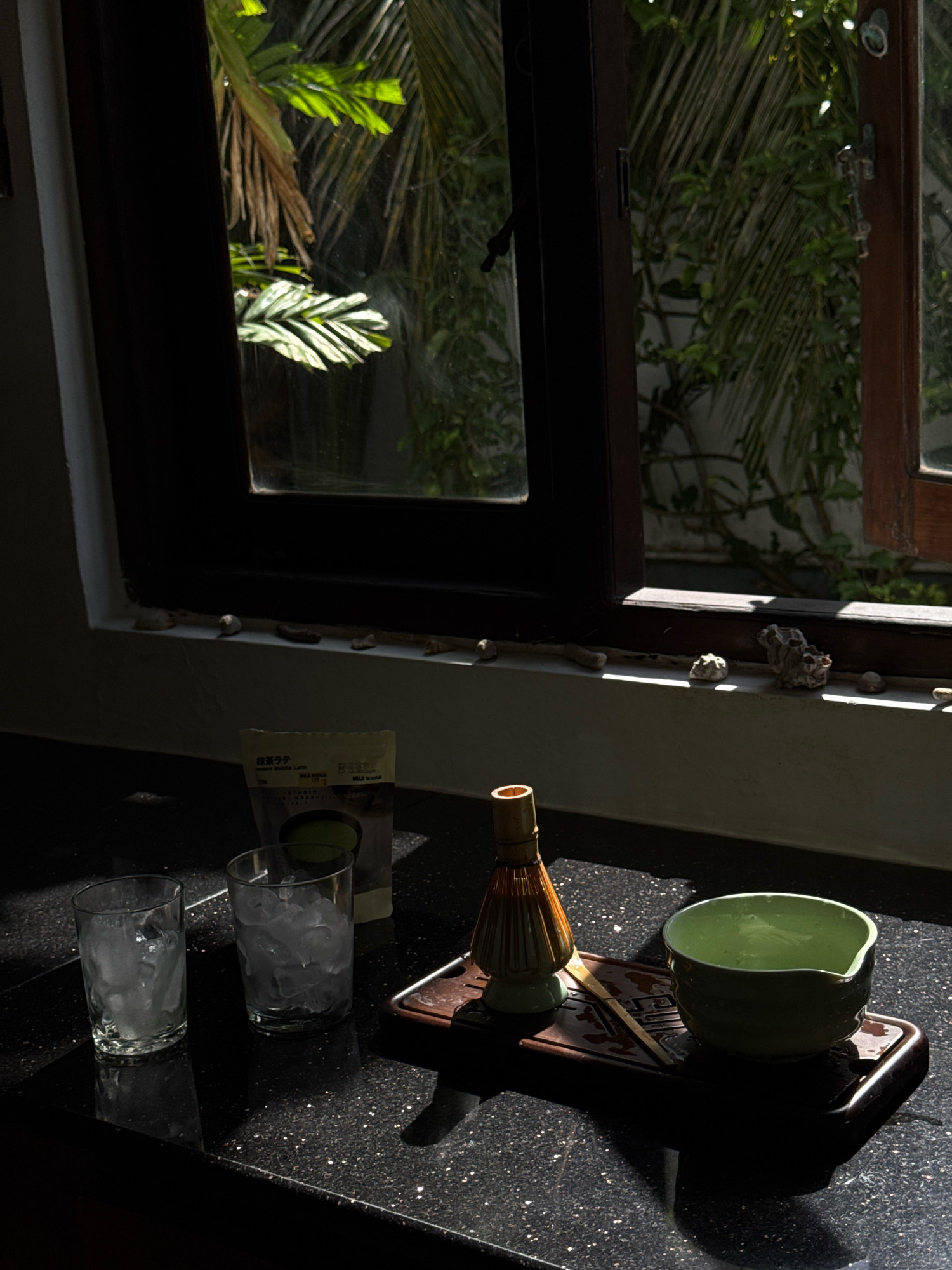 This is a still life image featuring a matcha tea set arranged on a dark, speckled countertop. The scene appears to be indoors, possibly in a kitchen or a preparation area, with sunlight streaming in from a window. The window is open, revealing lush green foliage and palm fronds outside, suggesting a tropical or subtropical environment. The location is identified as Goviyapana, Sri Lanka.
In the foreground, two clear glasses filled with ice are positioned on the left side of the frame. To the right of the glasses, a small, light-colored packet with Japanese text, likely containing matcha latte mix, is visible. The central focus of the arrangement is a dark wooden tray holding a matcha whisk (chasen), a bamboo scoop (chashaku), and a light green ceramic matcha bowl filled with liquid. The sunlight casts strong shadows across the countertop and the objects, creating a dramatic effect. The overall atmosphere is one of serene preparation for making matcha tea in a bright, natural setting. There are no people present in the image.