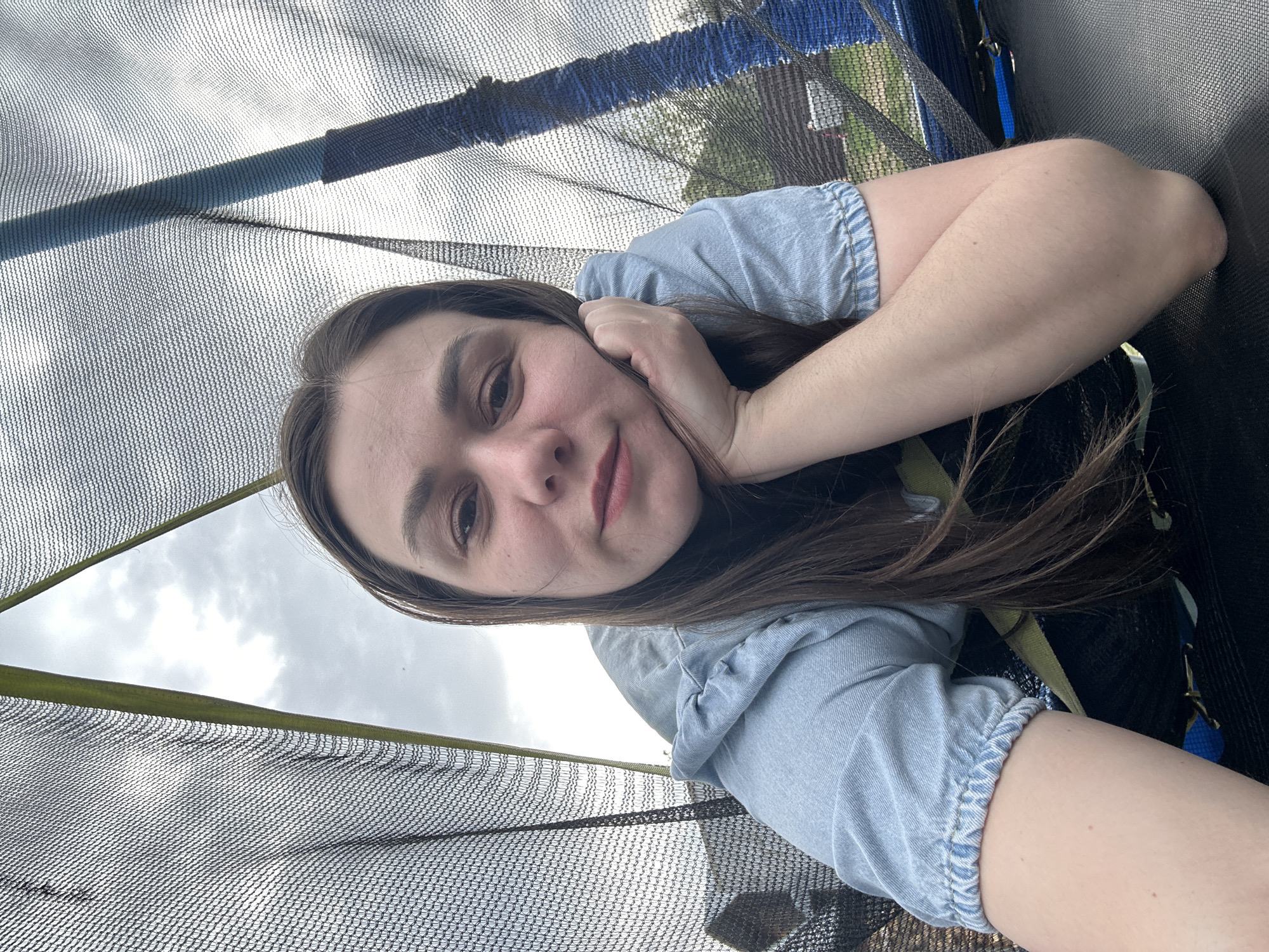 A young woman is taking a selfie while lying down inside a trampoline enclosure. She has long brown hair and is wearing a light blue denim jacket. The trampoline net surrounds her, and glimpses of the overcast sky and possibly a residential area are visible through the mesh. The location cannot be confirmed as Chaikovsky, Russia.