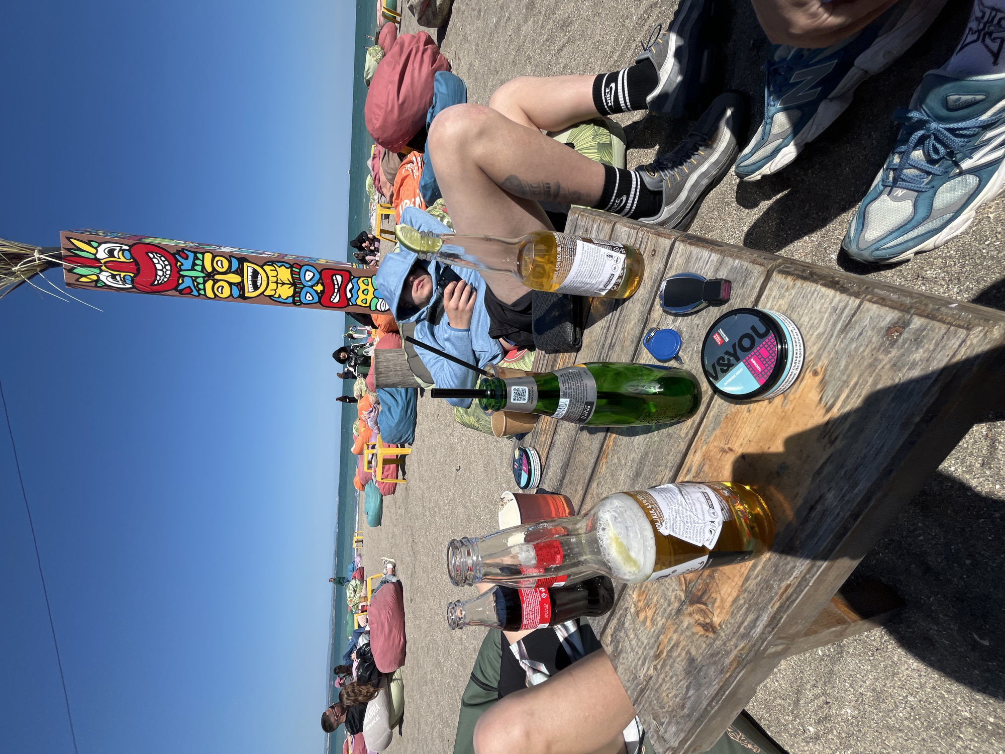 Here is a description of the image:
The image shows a sunny beach scene, likely during the day. The background features a blue sky and the Black Sea. In the foreground, there is a wooden table with various objects placed on it, including beer bottles (both empty and full), a Coca-Cola bottle, and a container of Swedish snus. At least two people are sitting at the table, visible through their legs and shoes. One individual is wearing black Nike socks and grey sneakers, while another has blue sneakers. There is also a person lying down next to the table wearing a blue hoodie and black shorts, presumably asleep or resting. Other people are relaxing on colorful bean bags and lounge chairs lined up along the beach. There is a colorful totem pole and beach-themed decorations, and colorful bean bags and lounge chairs aligned at the coast. This scene is in Burgas, Bulgaria.