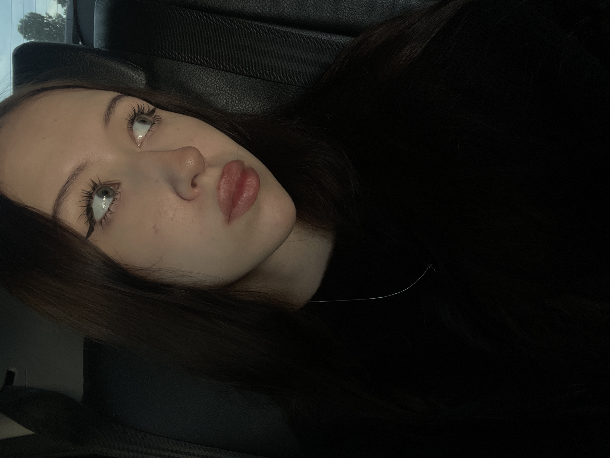 A young woman with long, dark hair and light-colored eyes is pictured indoors, likely inside a vehicle. She is wearing a black top and a thin silver necklace. Her makeup includes winged eyeliner and mascara. The background shows a dark leather seat and a portion of a seatbelt. The lighting is dim, suggesting it may be evening or nighttime. The image does not provide enough information to determine the specific location within Gavrilovka, Russia.