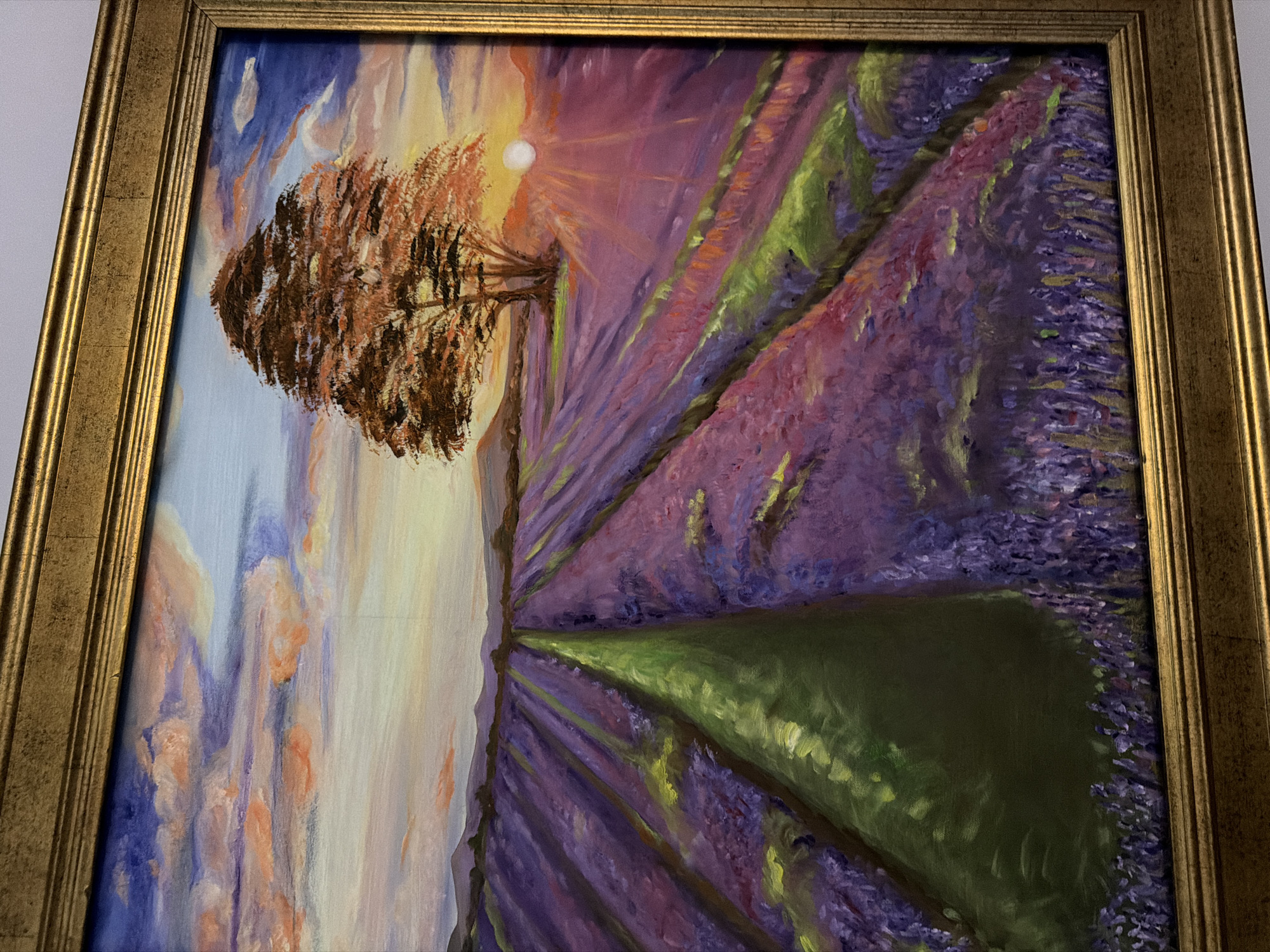 A framed oil painting depicts a sunset over a lavender field with a single tree and mountains in the background. The sky is filled with clouds painted in shades of blue, pink, and orange. The lavender rows are rendered in deep purples and pinks, with streaks of green indicating pathways or shadows. A solitary tree with reddish-brown foliage stands to the right of the frame. The sun, a bright white circle, is setting behind the mountains, casting rays of light across the landscape. The painting is in a gold-colored frame. There are no people or animals in the painting. The scene is a serene, idealized landscape. The image is not taken in Riviera Beach, United States of America.