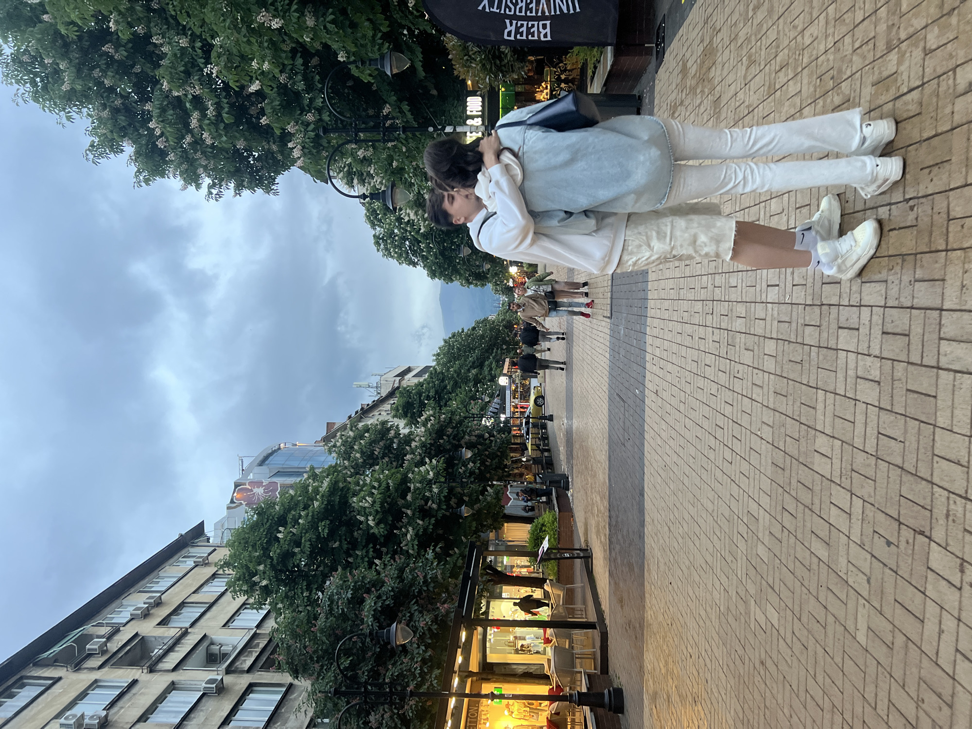 Two people embrace on a brick-paved sidewalk in Sofia, Bulgaria. The person on the left wears a white long-sleeved shirt and light-colored shorts, and white sneakers with black Nike logos on their socks. The person on the right wears a white long-sleeved shirt, light blue denim jacket, and white pants, and white sneakers. A black bag hangs on the person on the right. A banner, partially visible, reads "BEER UNIVERSITY." Other people walk further down the sidewalk. Trees line the sidewalk. Buildings are visible on the left, and streetlights are interspersed along the sidewalk. The sky is overcast.