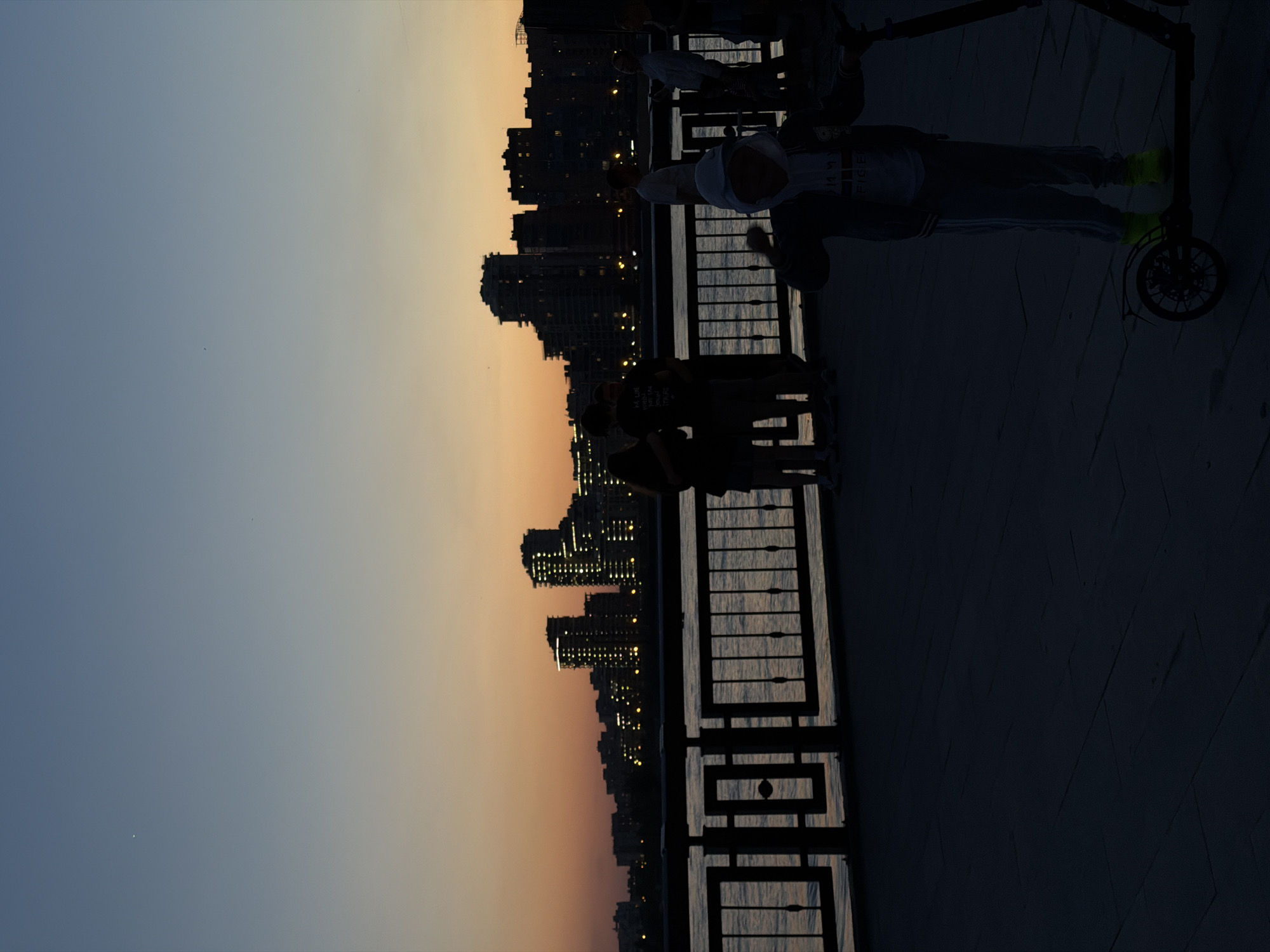 The image captures a twilight or dusk scene, likely in a city overlooking a body of water. On the right side, a dark foreground suggests a paved area with a decorative railing. Silhouetted figures of people stand and engage near the railing. A person is standing on a scooter. Across the water, a dense cityscape glows with the lights of buildings. The sky graduates from a light blue at the top left to a soft orange near the horizon. The scene suggests a waterfront promenade or viewpoint at twilight. The location may be Kazan, Russia given the prompt.
