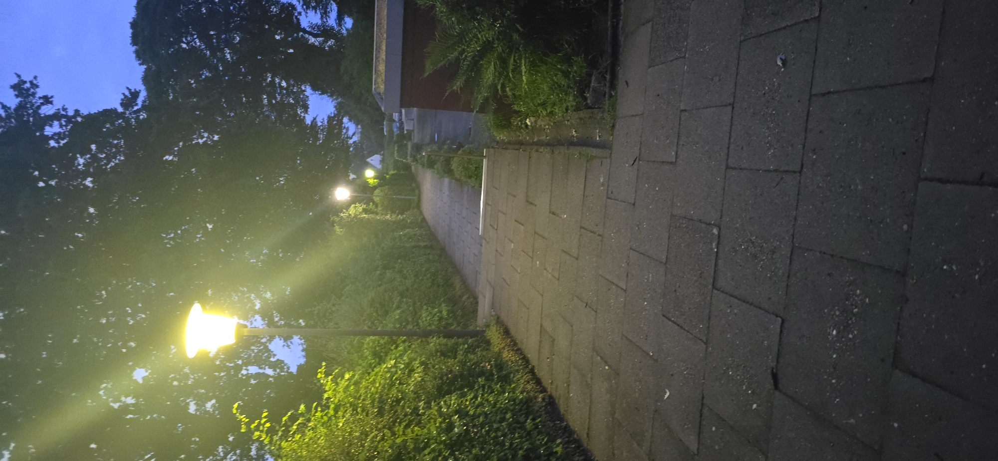 A dimly lit pathway made of dark grey concrete paving stones, some with a slightly lighter speckled pattern, leads away from the viewer. The path appears to slope gently downwards and to the right. It is bordered on the left by a dense, neatly trimmed hedge, illuminated by a tall, modern lamppost emitting a bright yellow light. The lamppost is dark grey and has a conical shade. Further down the path, to the right and slightly further away, another lamppost is visible, also emitting a yellow glow. Beyond the hedges and lampposts, the scene is dominated by the dark silhouettes of trees against a darkening blue sky. To the right of the path, a low, red-brick building with a dark, flat roof and dense green foliage in front of it is visible. Cars are parked in the distance beyond the building. The image appears to have been taken in the evening or early morning. The location is a residential area in Seevetal, Germany, likely a park or communal pathway within a housing estate.