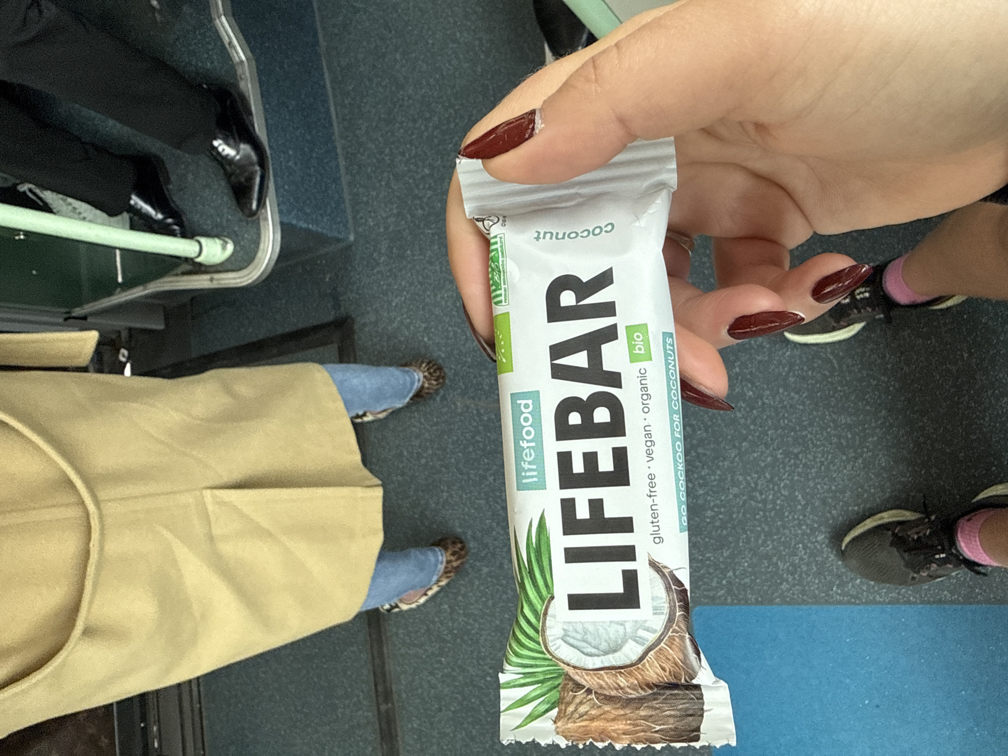 A first-person perspective photograph depicts a hand holding a "lifefood LIFEBAR coconut" snack bar. The background includes: seated person wearing a tan overcoat, jeans, and animal-print shoes; and standing person wearing black sneakers and pink socks. Additionally, there is a man's leg and shoe visible in the top left corner. The setting appears to be inside a train, potentially the Paris Metro, given the interior's color scheme and the presence of passenger seating.