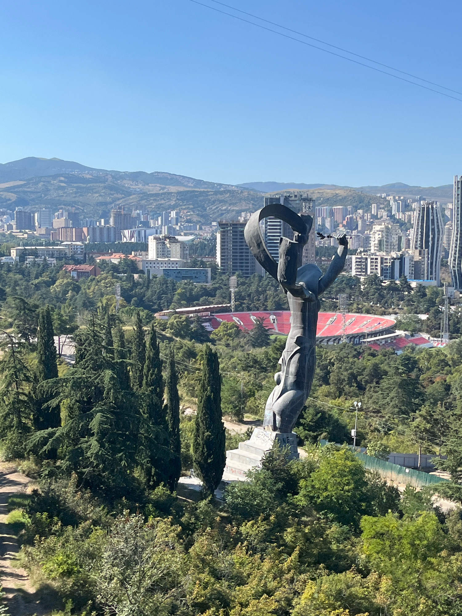 The image shows the "Mother of Georgia" statue in Tbilisi, Georgia, overlooking the city. The statue, a woman holding a sword and a bowl of wine, is made of bronze and stands on a hill. In the background, the cityscape of Tbilisi stretches out, with numerous buildings of various architectural styles, including modern skyscrapers and older structures. A red-seated stadium is visible amidst the greenery. The scene is set against a backdrop of rolling hills and a clear blue sky. The foreground is dominated by lush green trees, including tall cypress trees, and dense vegetation. A pathway is visible to the left of the statue.