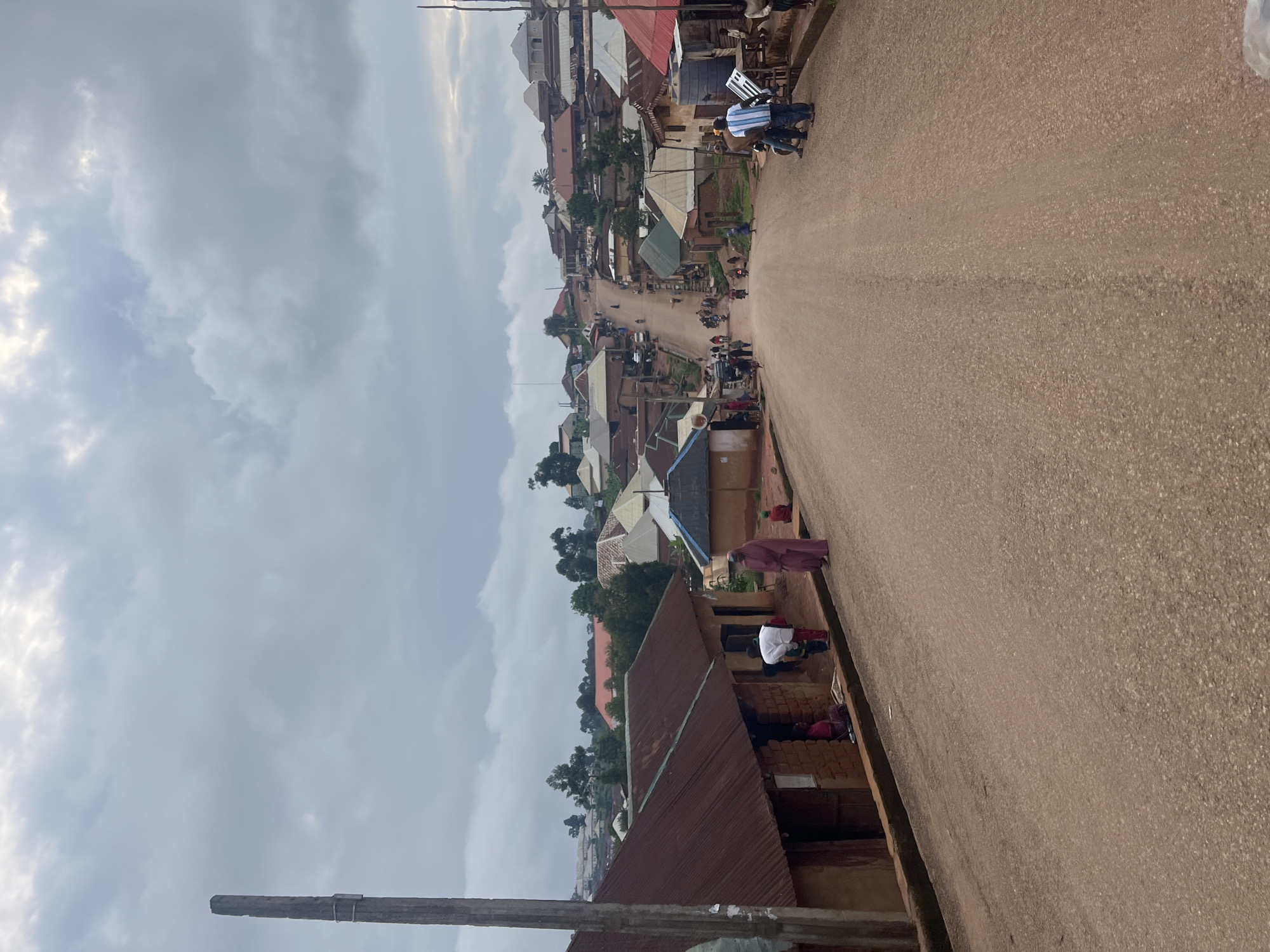 This image captures a street scene in Gembu, Nigeria, under a cloudy sky. The perspective is from a high angle, looking down a dusty, reddish-brown road lined with buildings. The architecture is varied, with many structures featuring corrugated metal roofs, some rusted and others appearing newer. Some buildings are multi-story, while others are single-story structures.
Several people are visible in the scene. On the right side of the road, near the foreground, a person in a maroon garment is standing, and further down, a person in a white shirt and red bottoms is kneeling or sitting. There are other individuals in the background, some on motorcycles and others walking. The presence of numerous motorcycles suggests a common mode of transportation in this area.
The overall atmosphere appears to be that of a typical day in a bustling community. The cloudy weather suggests it might be overcast, potentially before or after rain. The time of day is difficult to ascertain precisely, but the lighting doesn't indicate harsh sunlight. The scene depicts a lived-in environment, with everyday activities taking place on the street. There is no visible text in the image.