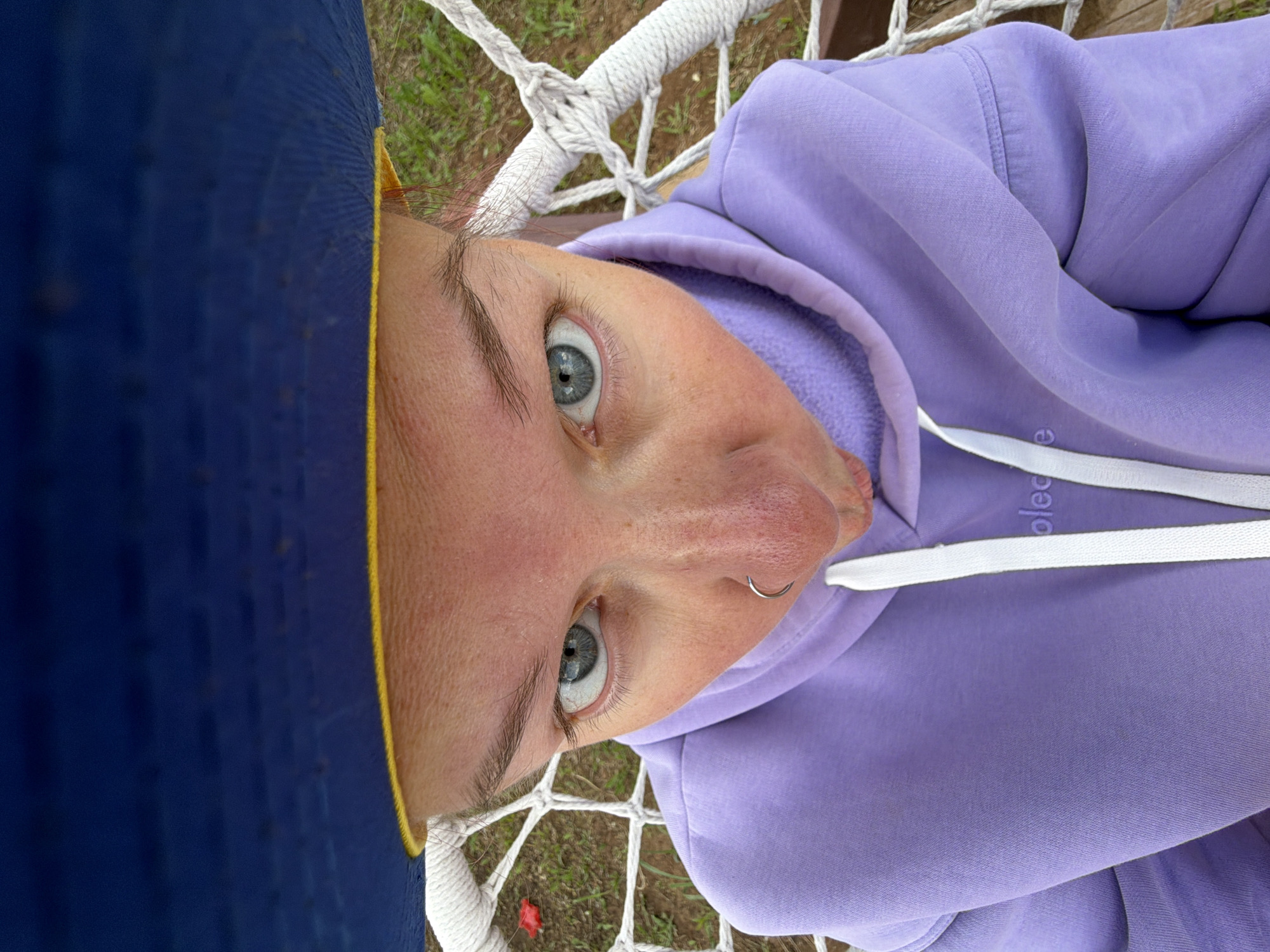 A close-up, high-angle portrait captures a person lying in a white rope hammock or net swing, looking directly upwards at the camera. The subject has light blue-grey eyes, prominent eyebrows, and a small silver hoop nose ring on their right nostril. Part of a dark blue cap or hat with a yellow rim is visible on the left side of the frame, partially obscuring the person's head.
The person is wearing a light lavender or purple hoodie. White drawstrings with plastic tips are visible on the hoodie, and faint text, appearing to be "oled" or "olec," is embossed on one of the drawstrings. The background, seen through the gaps in the white rope netting, shows green grass and some brown earth, indicating an outdoor setting. The lighting is natural and appears to be daytime. The scene suggests a relaxed and casual moment, likely in Chaikovsky, Russia.