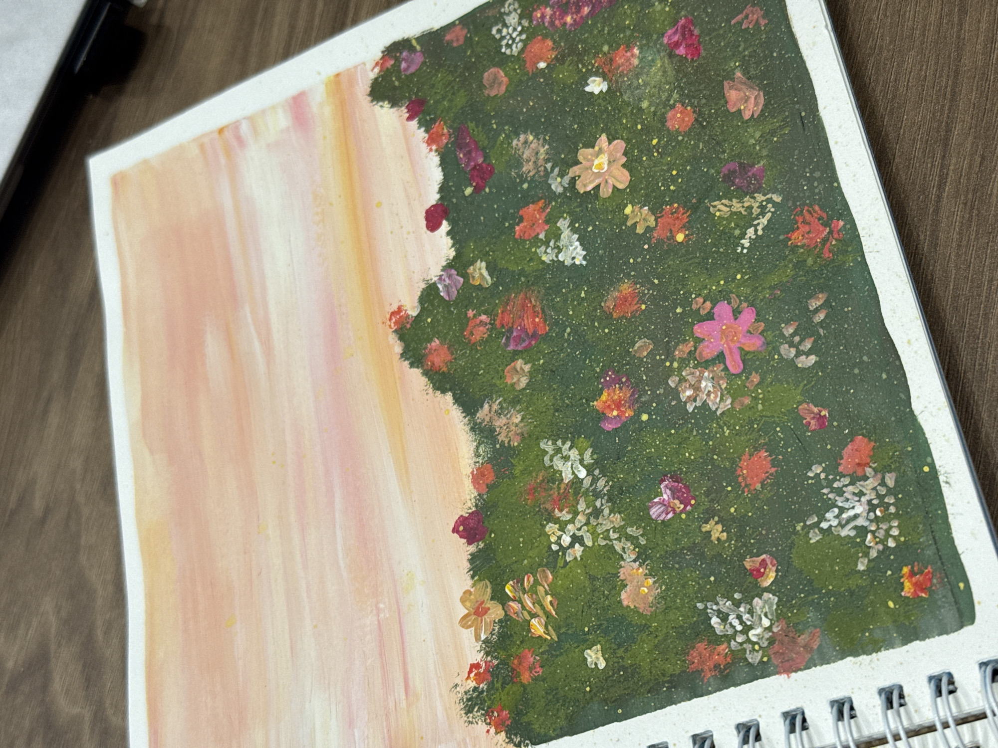 An aerial view of a painting on a wooden surface. The painting depicts a field of flowers against a sunset sky. The sky is rendered in soft strokes of pink, orange, and yellow, with a few scattered yellow dots suggesting starlight or distant light. Below the sky is a dark green, textured band representing a hillside or meadow, adorned with numerous small, impressionistic flowers in shades of red, pink, white, and yellow. The painting is bound in a white spiral notebook, the edge of which is visible on the left side of the frame. The wooden surface appears to be a table or desk. There are no people or animals visible in the image, and no specific location in the city of Murom, Russia, can be identified.