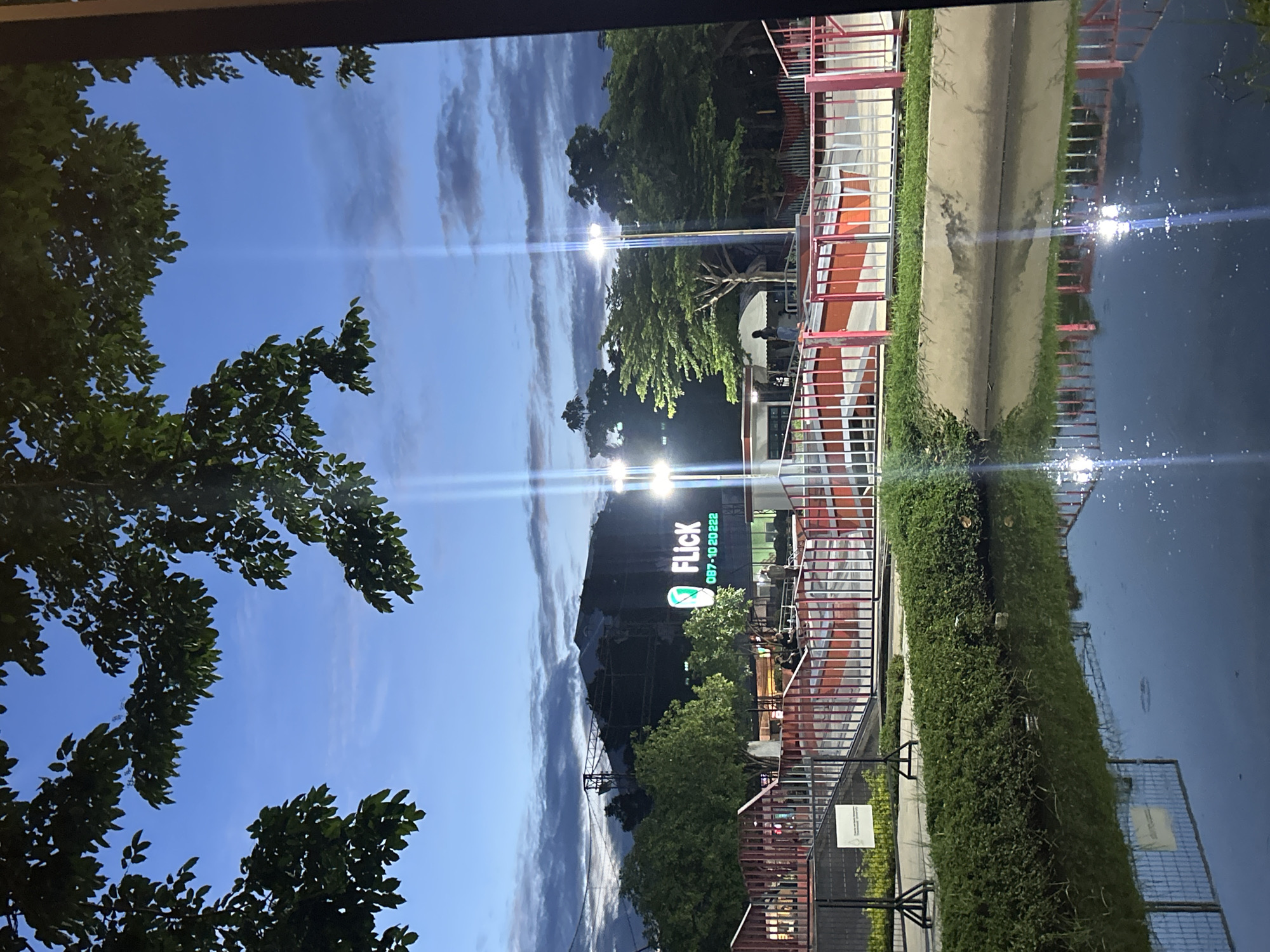 An outdoor scene at dusk features a skate park with red and white railings and ramps. A body of water with reflections of the sky and park is in the foreground. Behind the skate park, a building with a large illuminated sign reading "Flick" and a phone number is visible. Trees and streetlights are also present. The sky is a deep blue with scattered clouds. The location is Tha Khlong Town Municipality, Thailand.