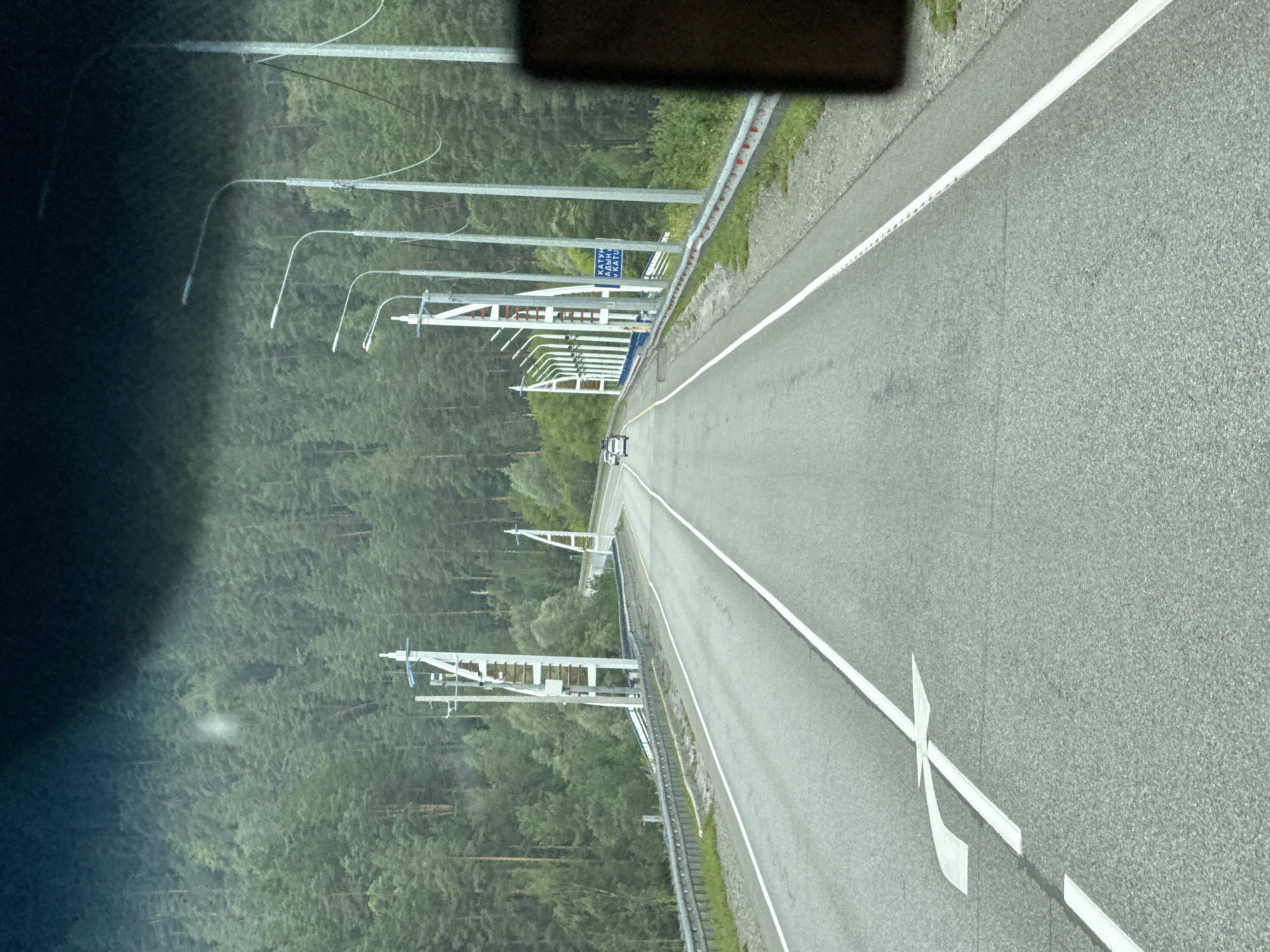 A paved road with two lanes and a white dividing line runs through a forested area. On the left side of the road is a metal guardrail. Along the road are multiple streetlights on tall poles and some large white structures that appear to be decorative or functional supports for overhead wires. Two white cars are visible driving away from the camera on the right lane. A blue sign with white lettering is mounted on a pole on the right side of the road. The surrounding environment is dense with tall, green coniferous trees. The road appears to be in a rural or mountainous region. The image seems to be taken from inside a vehicle, looking forward through the windshield. This appears to be the Katunsky Bridge area in Ust'-Sema, Russia, which is a common point of interest and traffic hub.