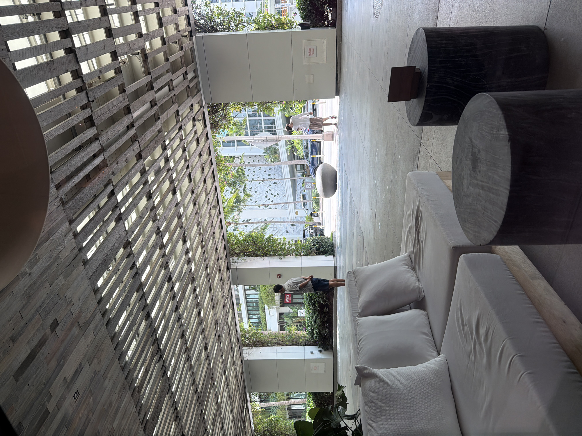 The media file displays an outdoor or semi-outdoor lounging area, likely a common space in a modern building in Miami Beach, United States of America.
**Subjects:** The scene features white outdoor lounge furniture, including a long sofa with multiple cushions, and two large, dark cylindrical tables or stools. On the left, a prominent architectural element consists of a slatted wooden ceiling or wall, possibly a pergola or sun screen, with light visible through its gaps. In the open background, lush tropical foliage, including palm trees and dense bushes, is visible. Two men are present: one is seen walking further into the distance on a paved surface, while another man stands closer to the foreground, facing away from the camera, near some greenery. Modern buildings with large windows are visible in the background. A dark car is parked on a street in the mid-ground.
**Activity or event:** The men appear to be casually traversing or standing in the outdoor space. There is no specific event or interaction clearly depicted. The scene suggests a relaxed atmosphere in a communal area.
**Notable details:** The time of day is bright daylight, indicated by clear lighting and shadows, suggesting a sunny day. The presence of palm trees and tropical greenery is consistent with the Miami Beach location. The architecture appears modern and airy, designed to integrate with the outdoors.
**Visible text:** The word "Avis" is clearly visible in red letters on a white sign in the background, near the standing man.