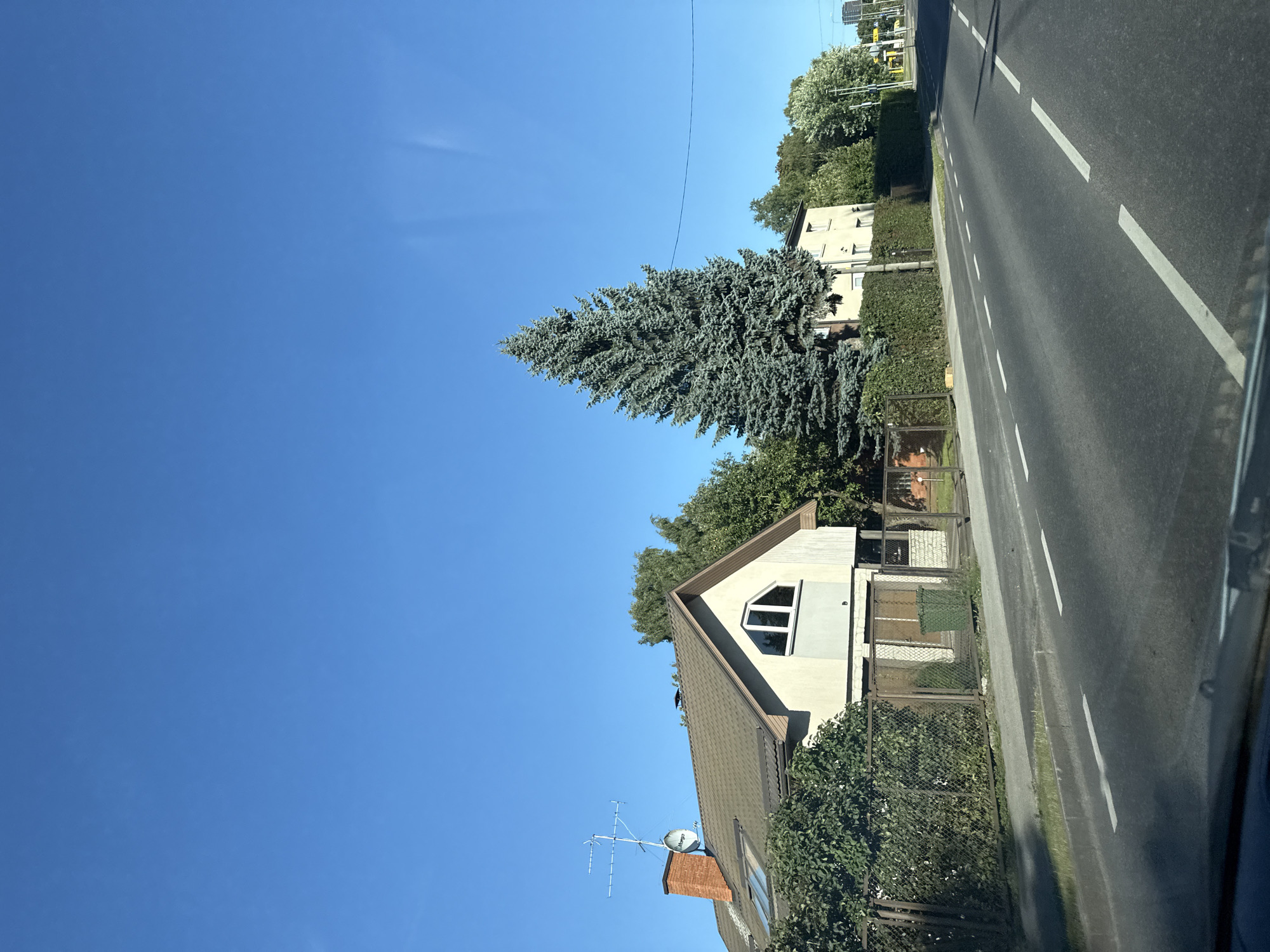 The image captures a view of a suburban street in Tallinn, Estonia, under a clear blue sky. On the left side of the road, a two-story house with a light-colored facade and a dark tiled roof is visible. It features a prominent brick chimney and a satellite dish. Adjacent to the house, a large, mature blue spruce tree stands tall. The property is enclosed by a metal fence and hedges. The road in the foreground has clear white lane markings, indicating it is a well-maintained thoroughfare. Further down the street, other buildings and trees are discernible, suggesting a residential area. The overall impression is one of a calm, sunny day in a quiet part of the city.