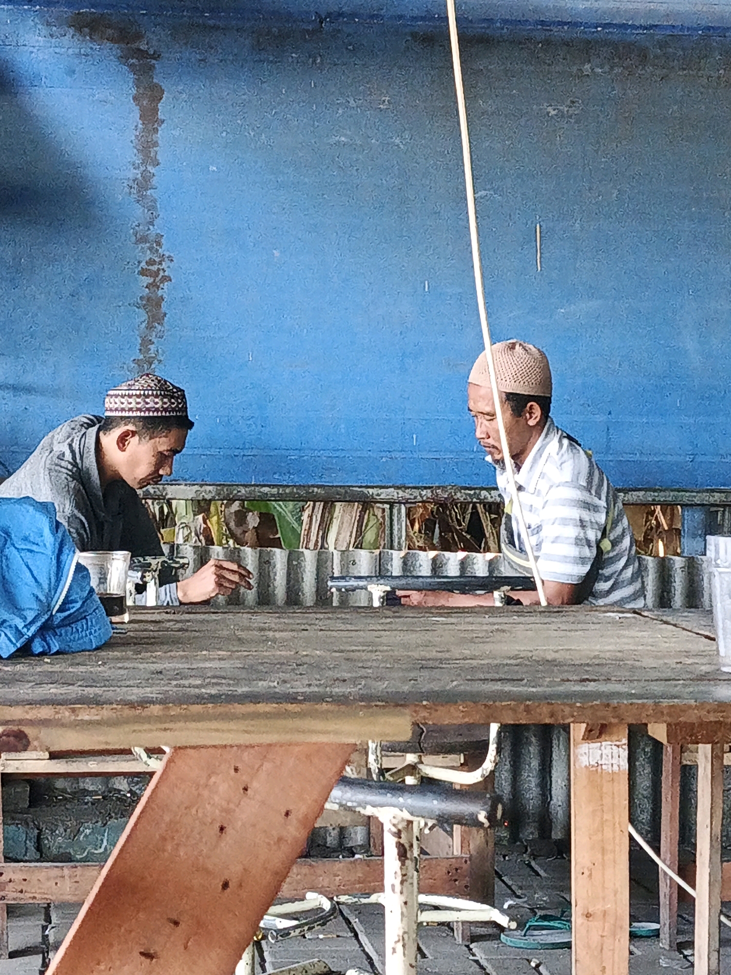 In the city of Ketegan, Indonesia, two men are seated at a wooden table in an open area with a blue corrugated metal wall behind them. The man on the left, wearing a gray jacket over a darker shirt and a patterned skullcap, leans forward, his right hand extended towards the table. A glass with a dark liquid is in front of him. The man on the right, dressed in a horizontally striped shirt and a tan skullcap, is looking down, his arms resting on the table. A thin white string hangs vertically in front of him, originating from the top of the frame. The table is weathered and appears to be a rough-hewn surface. In the background, more corrugated metal and glimpses of what might be foliage or debris are visible. The overall scene suggests a casual, outdoor setting, possibly a workshop or a resting area.
