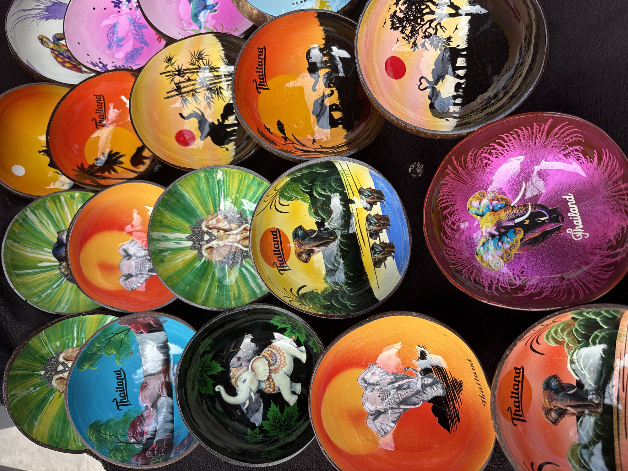 The image shows a collection of painted coconut shell bowls, likely for sale. Most bowls feature elephant designs and the word "Thailand," suggesting they are souvenirs. The colors vary, including orange, yellow, green, pink, and blue. They appear to be arranged on a dark surface, possibly a table or cloth, in a marketplace in Baan Bo Phut, Thailand.
