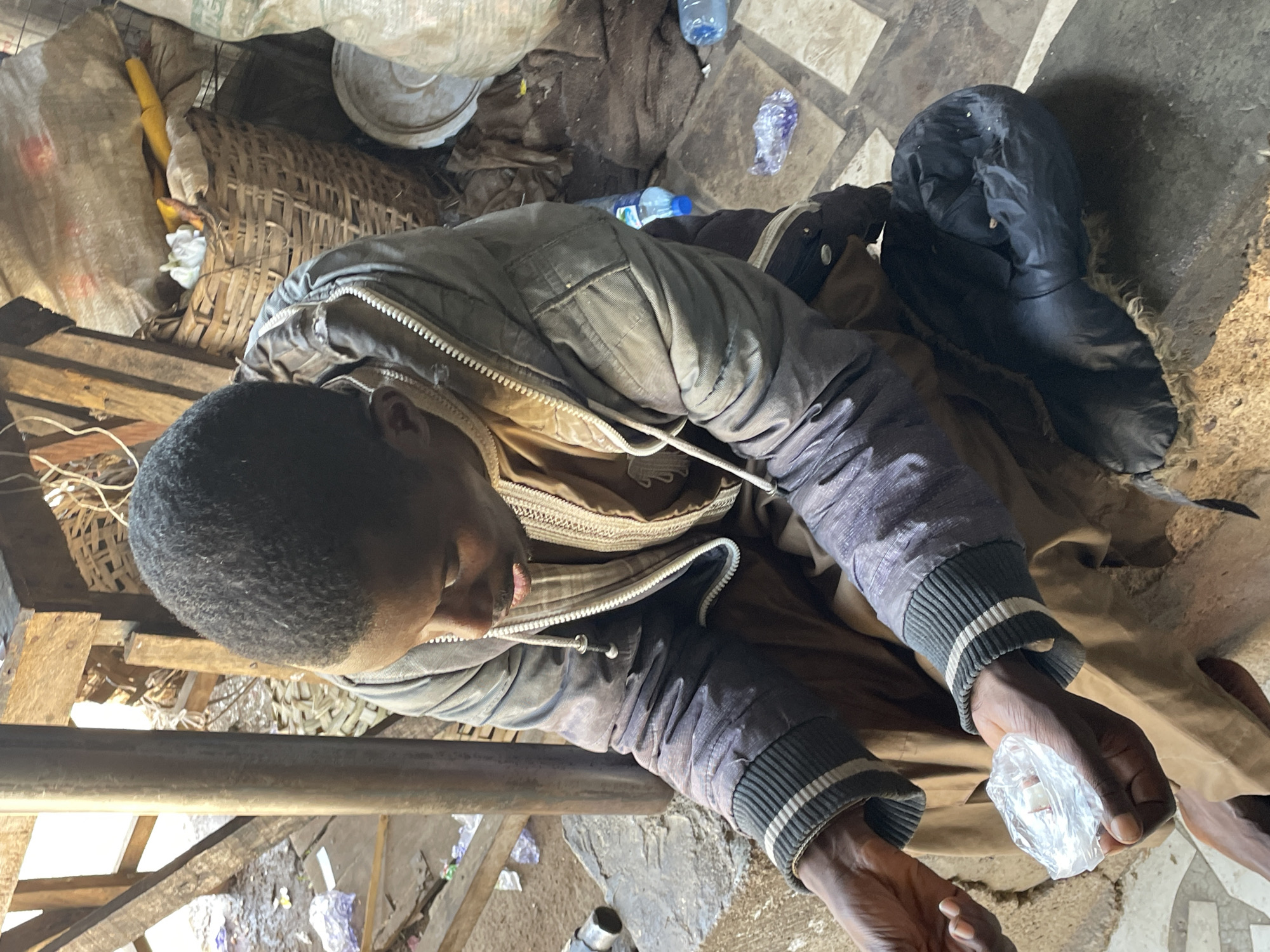 A person is lying in a cluttered environment. The person, of African descent, is clad in a jacket and appears to be holding a small plastic bag. The environment is littered with debris, including plastic bottles and trash. A basket and other containers are visible in the background. The scene appears to be indoors, though the exact location within Jos, Nigeria cannot be determined from the image alone.
